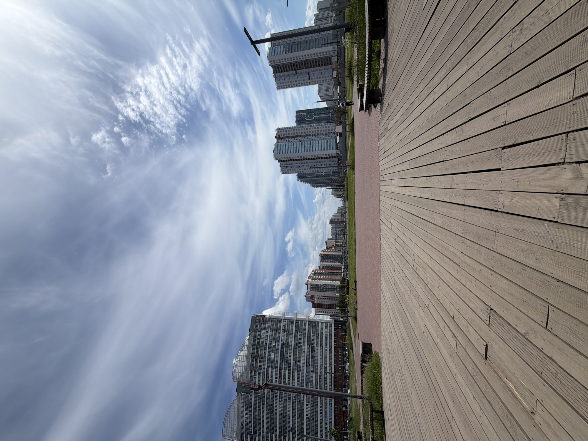 This wide-angle image captures a modern urban landscape in the Krasnoselsky District of Saint Petersburg, Russia, specifically within the Baltic Pearl residential area. The scene is observed from a low perspective, emphasizing a spacious foreground.
The upper two-fifths of the frame are occupied by a bright blue sky filled with a diverse array of clouds, ranging from wispy cirrus streaks to more defined cirrocumulus and cumulus formations, indicating a partly sunny day.
The foreground is dominated by a light brown wooden deck, composed of parallel planks receding into the midground. The planks exhibit minor wear and irregular spacing. On either side of this deck, partially visible, are sections of wooden benches positioned on unkempt green grass.
Beyond the wooden deck, a broad pathway or plaza of reddish-brown pavers extends towards the background. Flanking this paved area are strips of green grass, separating it from the buildings lining a wide avenue or street.
On the left side of the frame, a large, multi-story residential building features a distinct curved, arch-like structure over its upper section, likely composed of glass and metal. Its facade is light-colored with numerous windows. Further down the street, additional multi-story residential buildings are visible. Two yellow electric scooters are parked on the paved area near the first building.
On the right side, multiple tall, contemporary residential buildings with light-colored exteriors and blue-tinted windows line the street, receding into the distance. A prominent, dark-colored lamppost stands in the mid-right foreground. A single individual, dressed in light-colored clothing, is visible walking away from the viewer on the paved path towards the distant buildings.
The overall environment suggests a planned residential development with a focus on open spaces and modern architecture.
