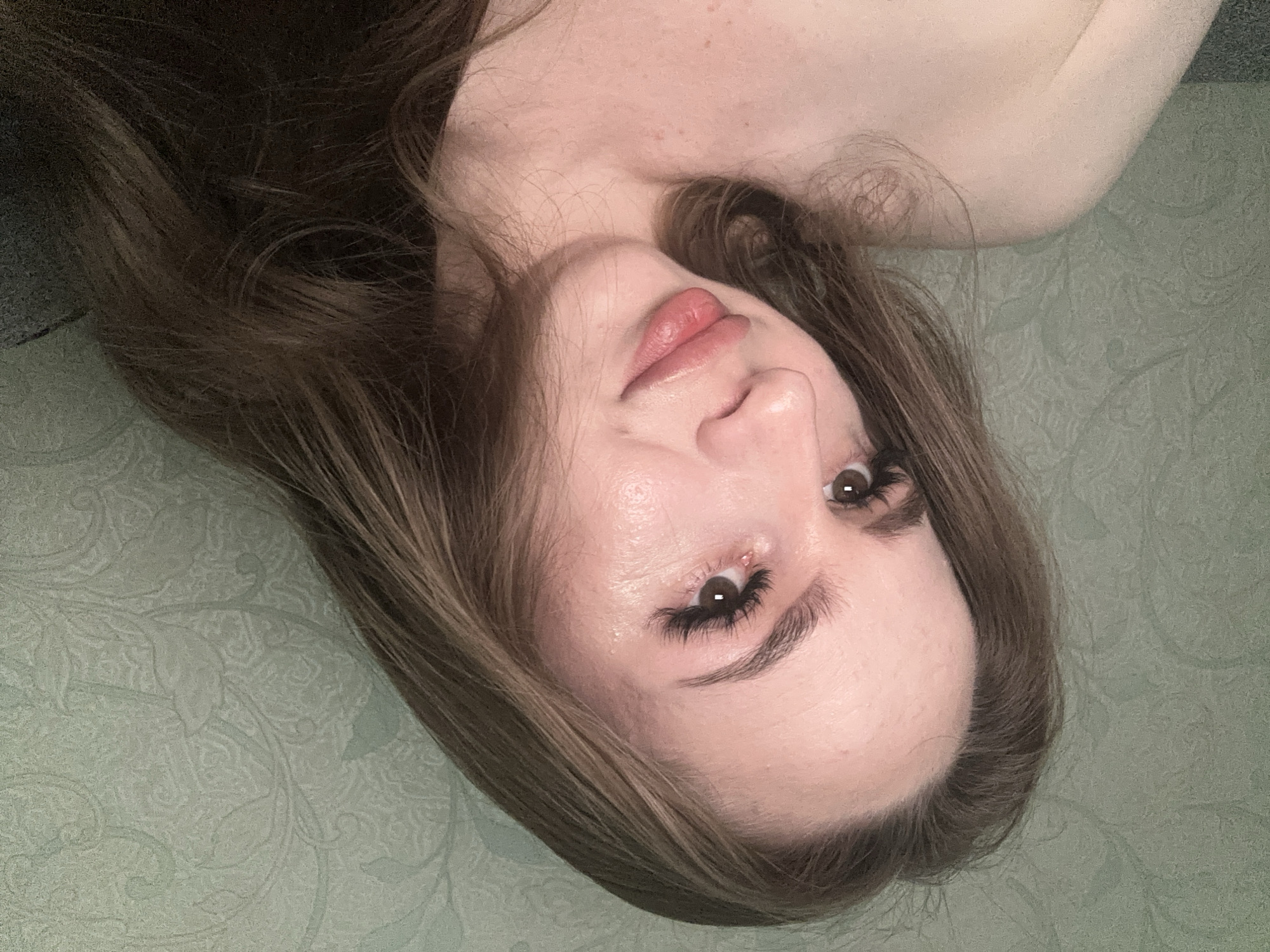 A close-up portrait features a young adult female from the chest up, positioned with her head slightly tilted and her gaze directed to the viewer's left, exhibiting a soft, subtle smile. She has long, wavy dark brown hair falling around her bare shoulders and upper chest. Her face shows brown eyes with pronounced dark eyelashes, well-defined eyebrows, a distinct nose, and full, light pink lips that are slightly parted. Her fair skin exhibits a subtle sheen on the forehead and cheeks, along with visible texture and scattered freckles or moles on her décolletage. The background is a wall covered with muted green wallpaper featuring an embossed or printed subtle, swirling botanical pattern. A dark, textured surface, possibly upholstery, is visible in the lower left and right corners. The location is Unknown, Unknown.