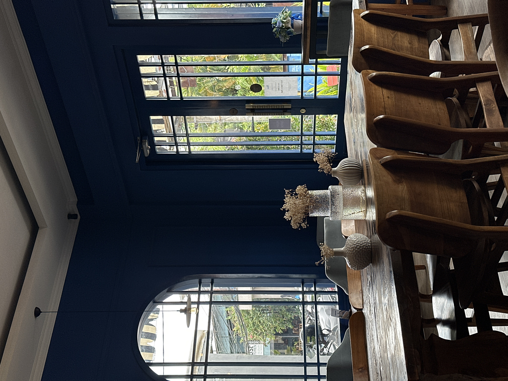 Captured from within a café or restaurant in North Kuta, Indonesia, the image reveals an interior setting characterized by blue walls. The focus is on a wooden table, around which chairs are arranged. Visible on the table are decorative vases filled with dried flowers, suggesting a deliberate attempt to create an inviting ambiance. Natural light streams through the gridded windows, providing views of the outdoors, indicative of a daylit scene within the café or restaurant. No people are visible in the image.