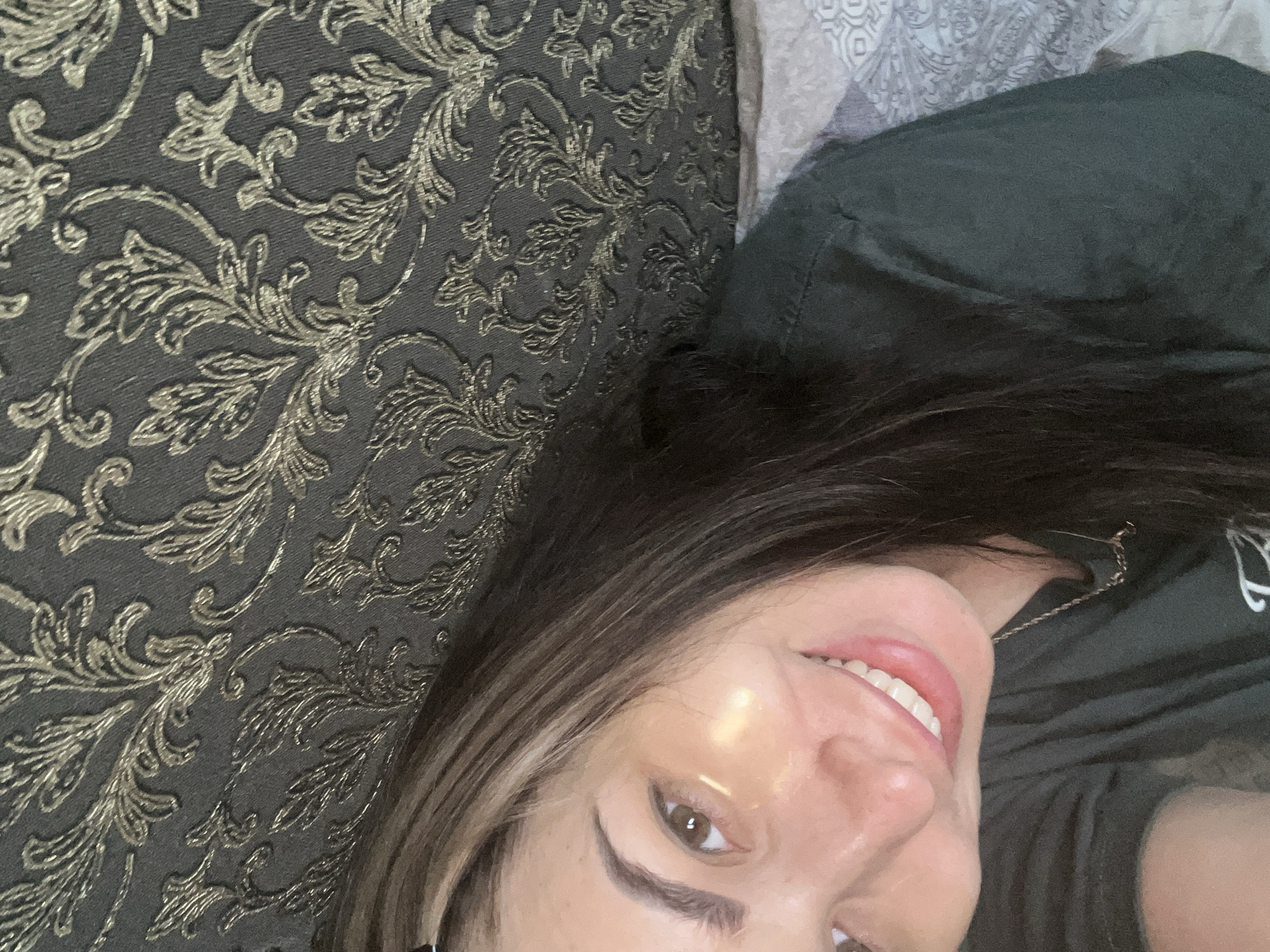 An angled, close-up shot captures a person, likely female, lying down and smiling broadly at the camera. Her head is tilted, with her face occupying the lower right portion of the frame. She has long, dark brown hair with lighter streaks, framing her face, and a warm skin tone with a subtle sheen on her forehead and cheek. Her right eye is open and looking directly at the viewer, and her wide smile reveals white teeth. She is wearing a dark green or olive-colored casual top, with a thin, gold-colored chain visible near her neck.
The background is dominated by a dark gray or charcoal-colored upholstered surface, possibly a sofa or bedding, featuring a rich, repeating metallic gold baroque or damask pattern. In the upper right corner, behind her head, a lighter-colored fabric with a subtle, possibly geometric or paisley design, is visible, suggesting another piece of bedding or furniture. The lighting appears soft, highlighting her features and the texture of the patterned fabric. No text is visible in the image.