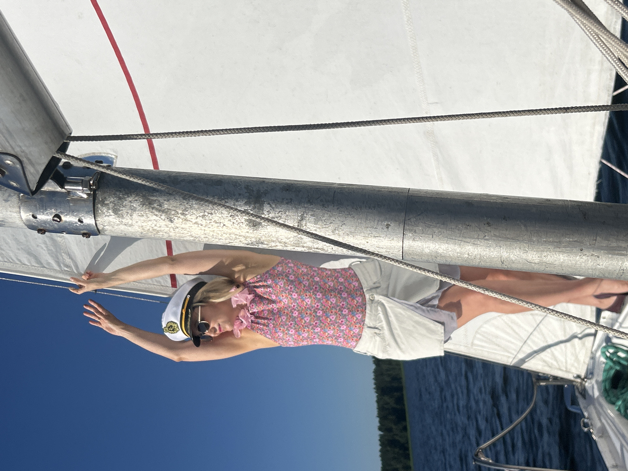 A woman wearing a captain's hat, sunglasses, and a floral-patterned halter top stands on a sailboat with her arms raised. She is wearing a light-colored skirt. The boat is on a body of water with a treeline visible in the distance under a clear blue sky. The mast and sail of the boat are prominent in the foreground. There is no information in the image to suggest this is located in Izhevsk, Russia.