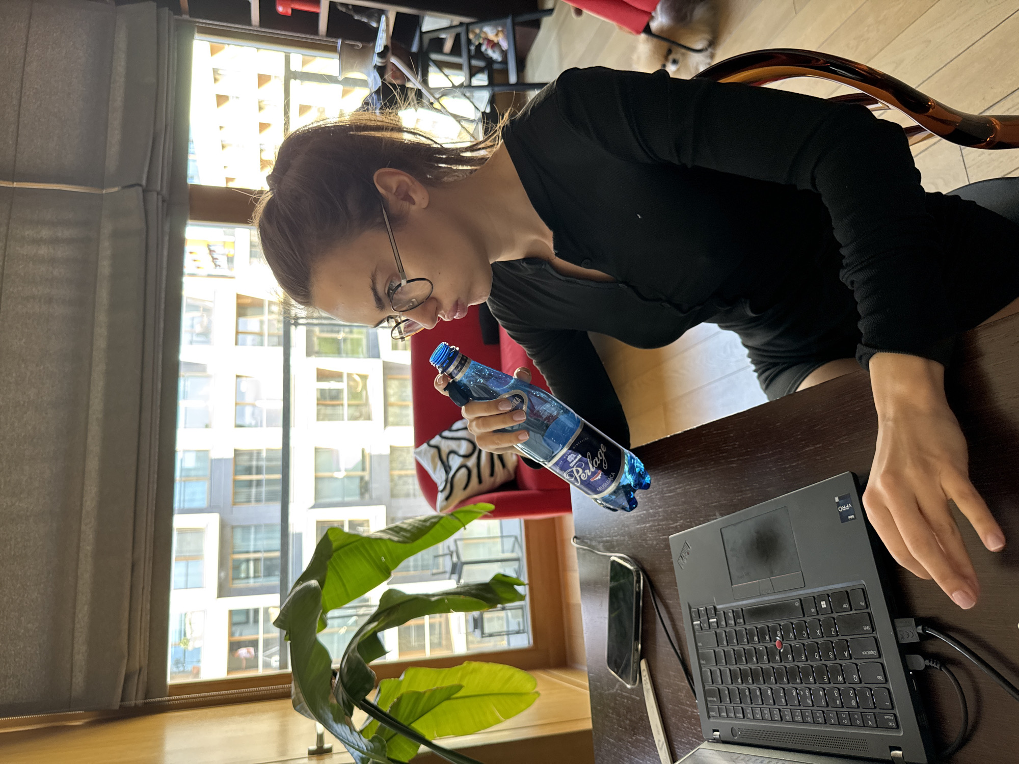 A woman with her hair pulled back, wearing glasses and a long-sleeved black top, sits at a dark wooden desk. She is holding a blue bottle of Perlage mineral water. On the desk is a black Lenovo laptop, a smartphone, and a stylus pen. A large green plant is visible in the lower left of the frame, and a red chair with a patterned white cushion sits behind the woman. Through the window behind her, modern apartment buildings are visible, suggesting an urban setting. This is likely in Warsaw, Poland, as the Perlage mineral water is a Polish brand.