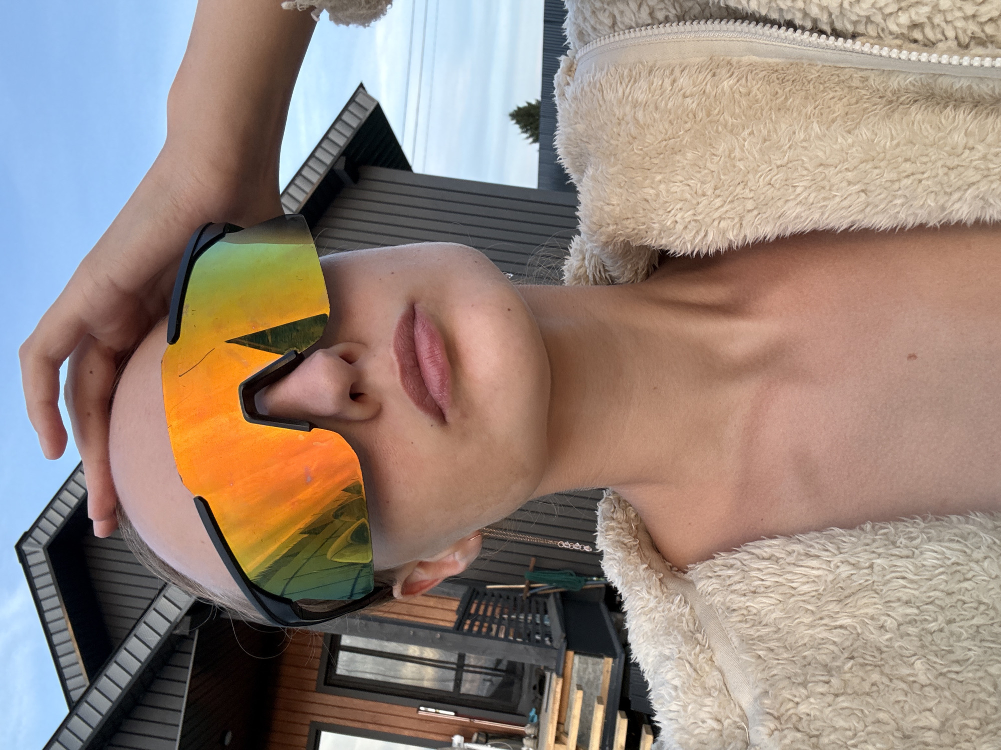 A close-up, vertical, eye-level shot shows a person's face and upper body, tilted slightly to the left, against a backdrop of a modern house and a clear sky. The person is wearing large, reflective sunglasses with orange and green lenses that mirror the surroundings. Their skin is fair, and their lips are slightly parted, painted with a matte pink lipstick. A fluffy, cream-colored jacket is visible, zipped up at the collar. The person's left arm is raised, with fingers gently touching their forehead or the top of the sunglasses.
The house features dark siding and a steeply pitched roof, with visible window frames. In the background, a small evergreen tree stands against the sky. The lighting suggests it is daytime, with a bright, clear sky, possibly indicating good weather. The overall impression is casual and relaxed, with the subject possibly posing for a selfie. There is no visible text in the image. The setting appears to be an outdoor residential area, possibly in a more rural or suburban part of Russia, given the location context.