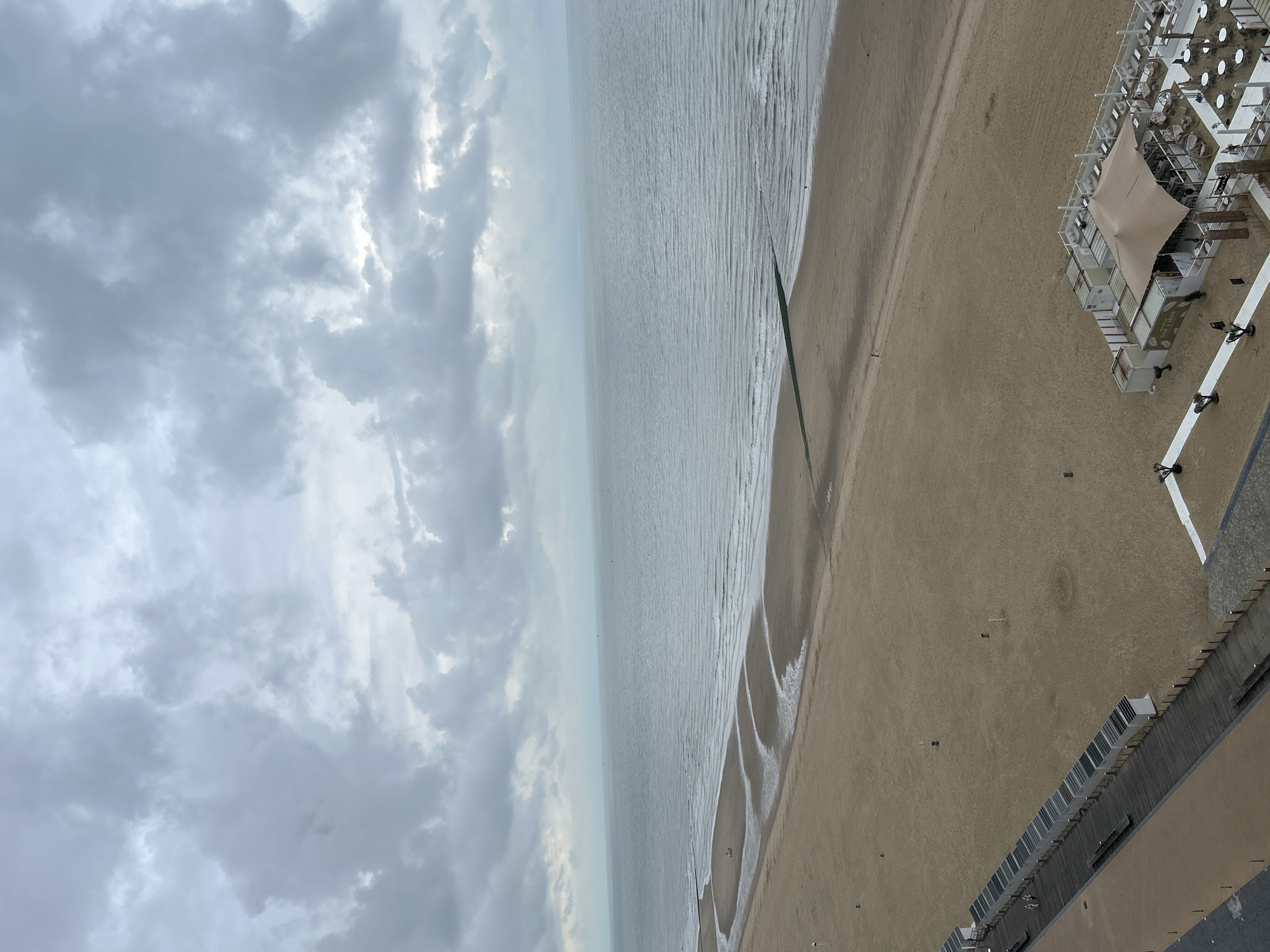 This is an aerial, wide-angle shot of a beach in Ostend, Belgium, on a cloudy day. The sea stretches to the horizon, with small waves lapping at the sandy shore. The beach is mostly empty, with a few scattered beach huts and a small beach bar with white seating areas and a beige tent. A wooden walkway runs along the edge of the beach, with two figures walking on it. A green barrier extends into the sea from the beach. The sky is filled with grey clouds, suggesting overcast weather.