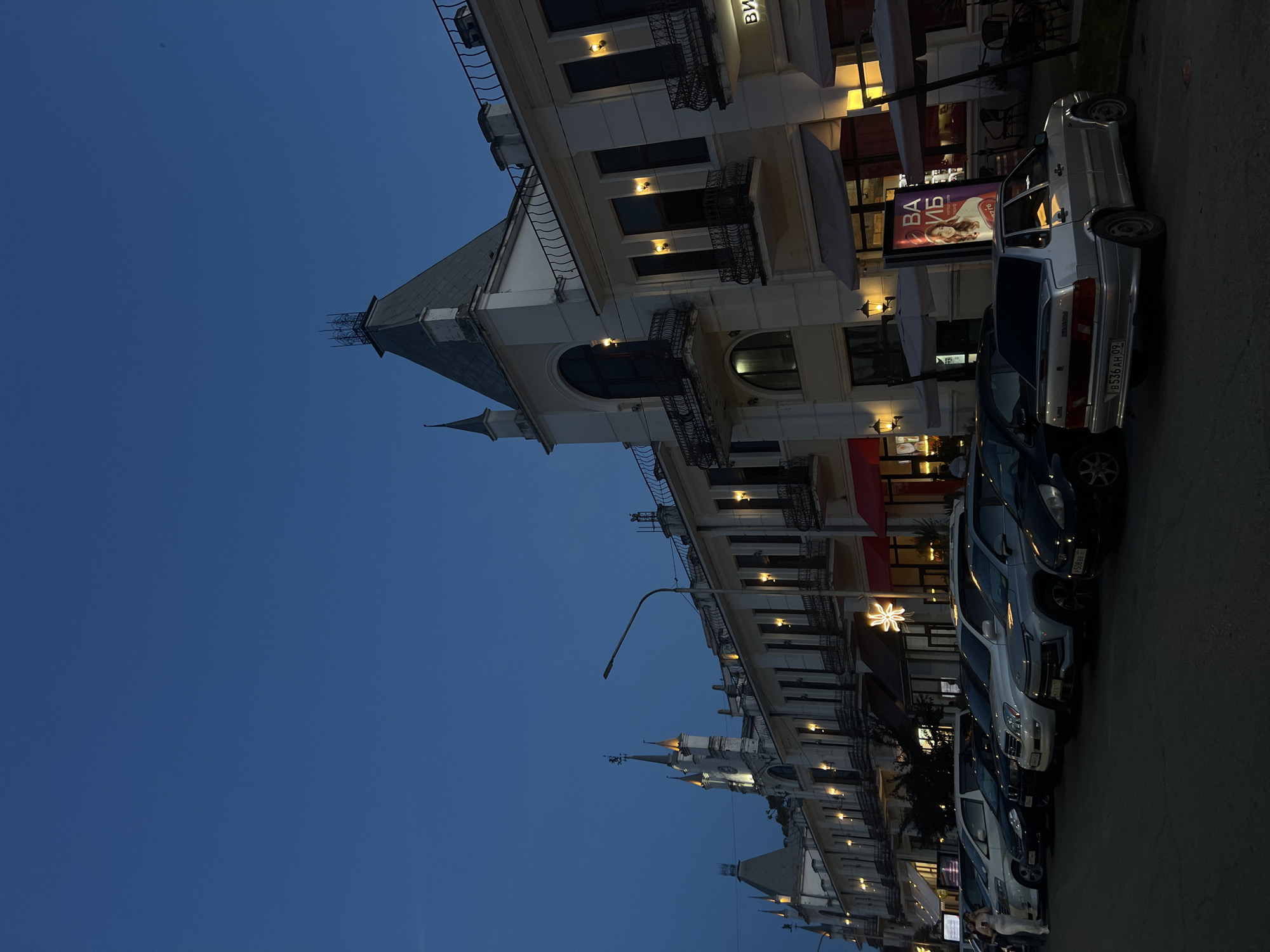 A low-angle, full shot captures an evening street scene in an unknown city, showcasing a row of parked cars on the left and a multi-story, white building with a gray, mansard roof and spire on the right. The building features multiple arched windows, balconies with black railings, and illuminated lamps. A street lamp stands in front of the building. Several cars, including a silver sedan, are parked on the asphalt street. A woman in a white dress is visible on the far left, standing near a white van. The sky is a deep, clear blue.