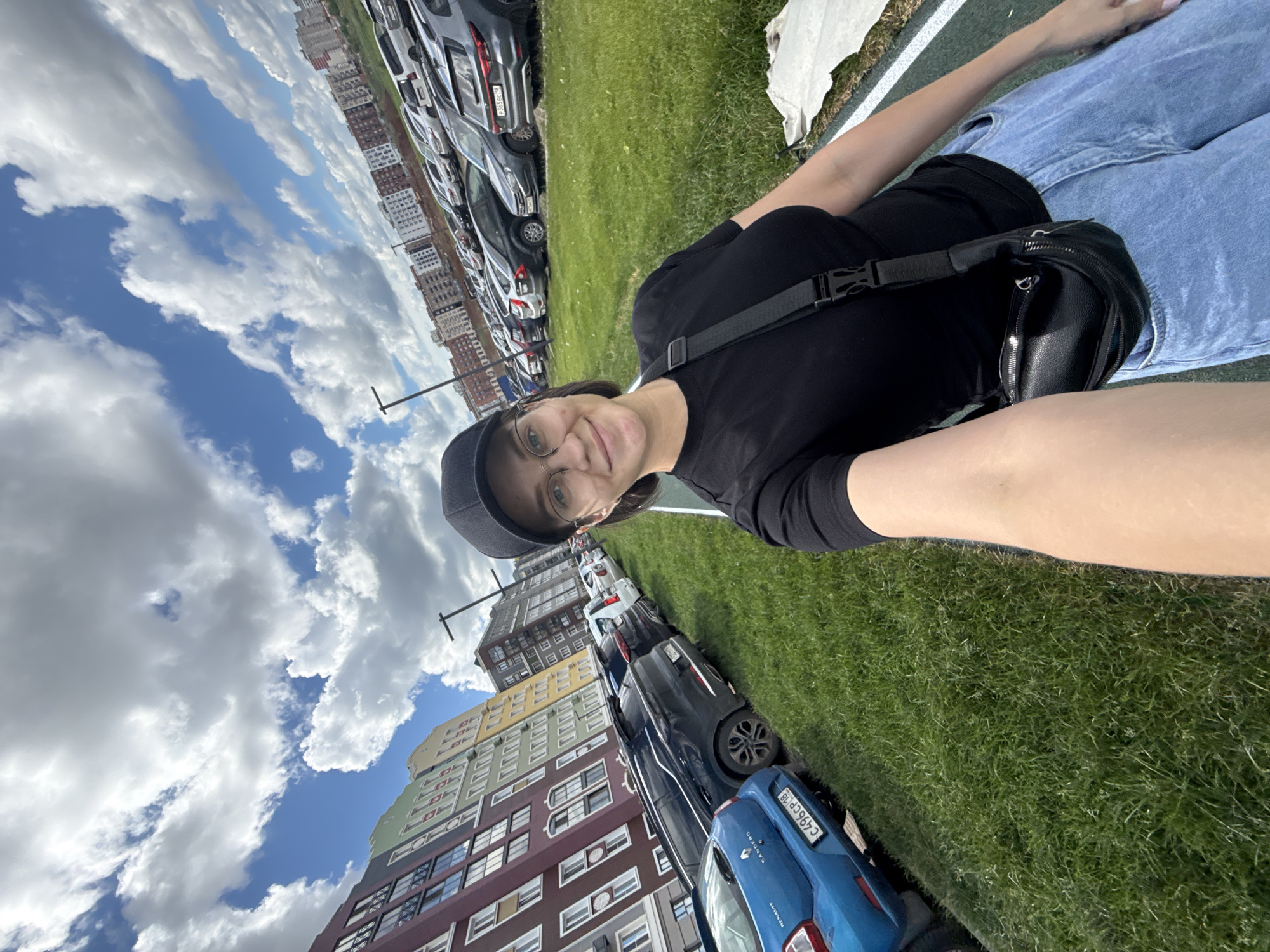 The image shows a young woman in a black t-shirt, blue jeans, a black baseball cap, and glasses, taking a selfie outdoors. She is wearing a black fanny pack across her chest. Behind her, a parking lot is visible, filled with cars. In the background, there are several multi-story residential buildings with colorful facades. The sky is partly cloudy. This scene appears to be in a modern residential area.