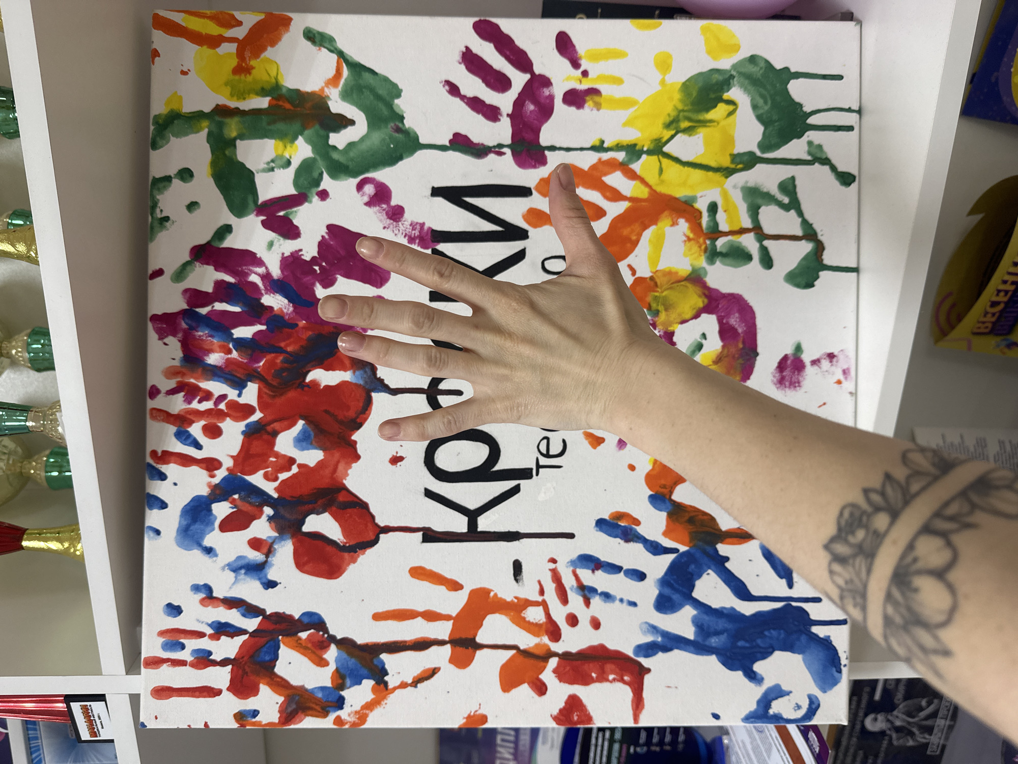 The image depicts a rectangular canvas with multicolored handprints in blue, red, orange, yellow, green, and purple on a white background. Black text is visible on the canvas. A human hand with pale skin is positioned over the center of the canvas. The lower arm of the person is visible, bearing a tattoo of a floral design.
The canvas is placed on a white shelf alongside various items. To the left, there are bottles wrapped in green and gold foil. To the right, there's a box with a blue and purple design.
The location of Votkinsk, Russia is not explicitly apparent from the provided image and its crops.