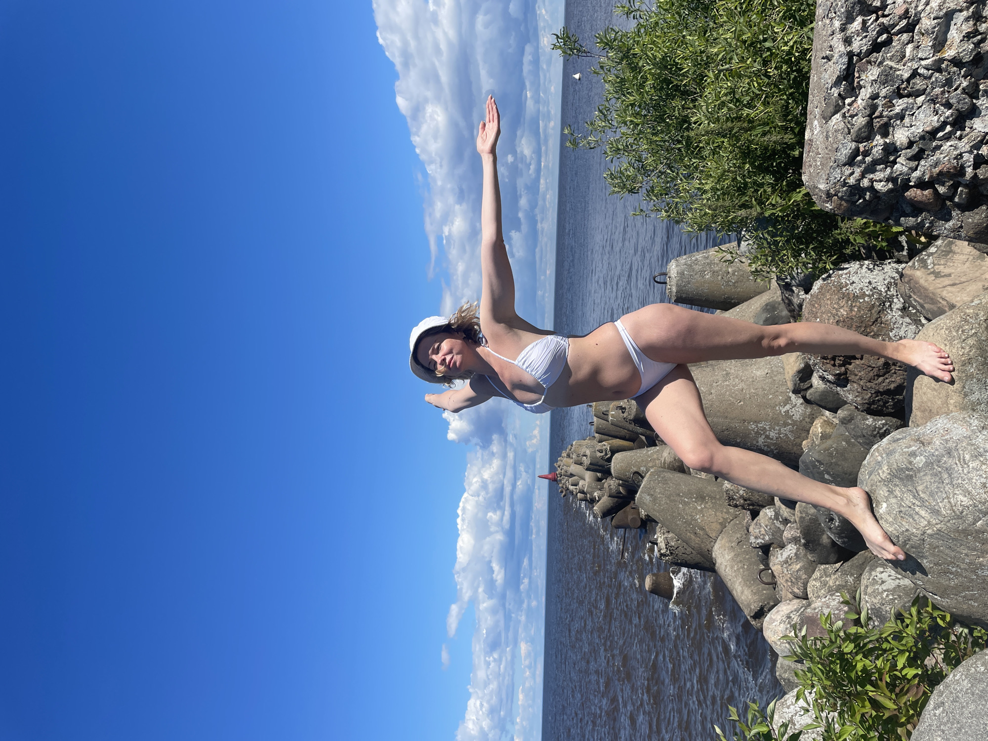 A blonde woman in a white bikini and white hat stands on a rocky breakwater with her arms outstretched. The breakwater is made of large concrete blocks and natural stones. The sea is dark and choppy, with a clear blue sky dotted with white clouds above. In the distance, a red buoy and some small white boats are visible. Lush green foliage grows on the right side of the breakwater. The scene appears to be the shoreline of Lake Beloye in Belozersk, Russia, known for its historic white-stone architecture and natural beauty.