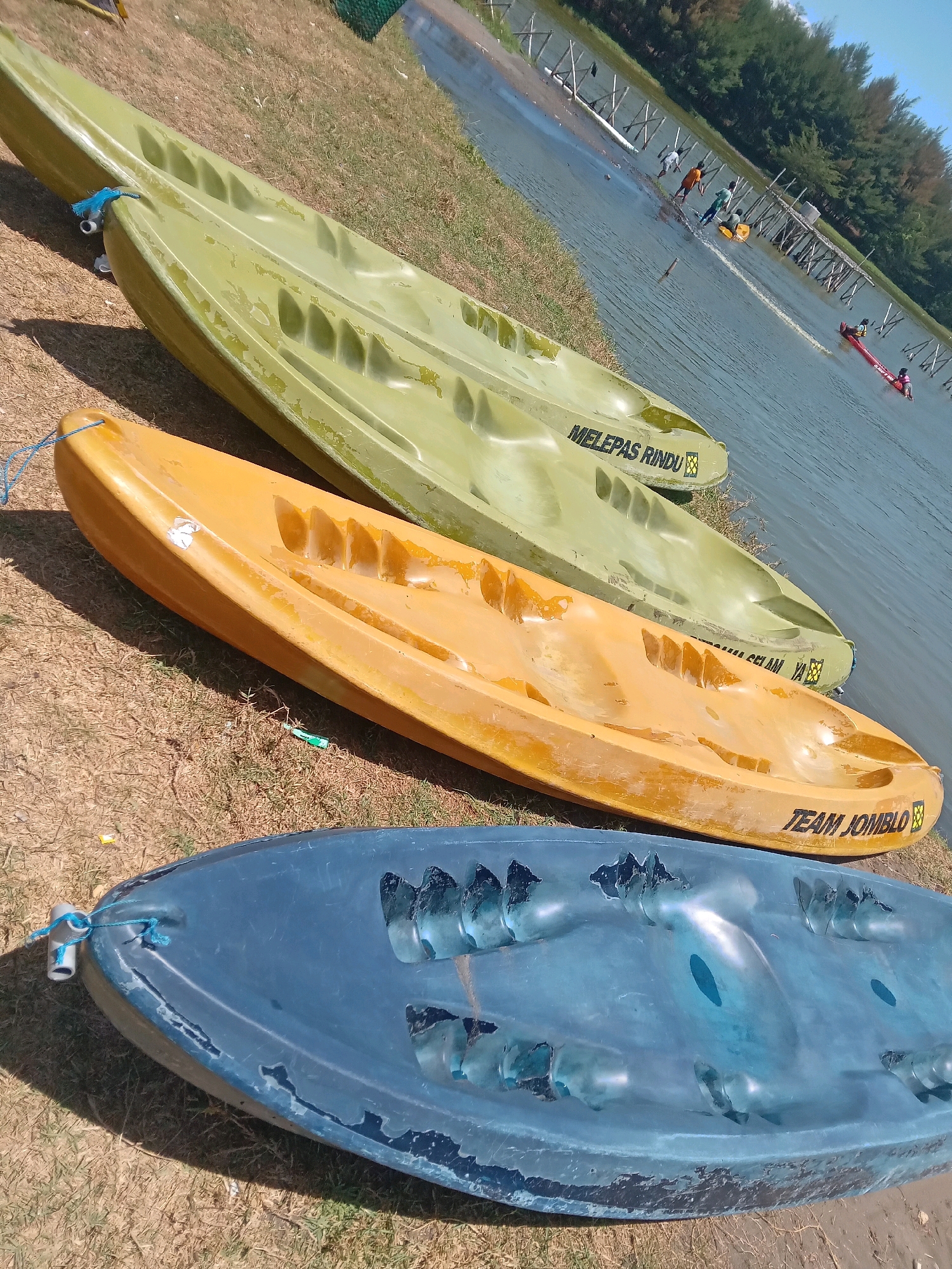 The image shows three kayaks lined up on a grassy shore next to a body of water. The kayaks are of different colors: the top one is green, the middle one is orange, and the bottom one is blue. Each kayak has text written on its side. The green kayak has "MELEPAS RINDO" written on it, and the orange kayak has "TEAM JOMBLO" written on it. In the water, several people are visible, some of whom appear to be participating in water activities. The background shows trees and some wooden structures along the shoreline. The overall scene suggests a recreational area, possibly at Glagah Beach in Indonesia, given the presence of the kayaks and the natural environment.