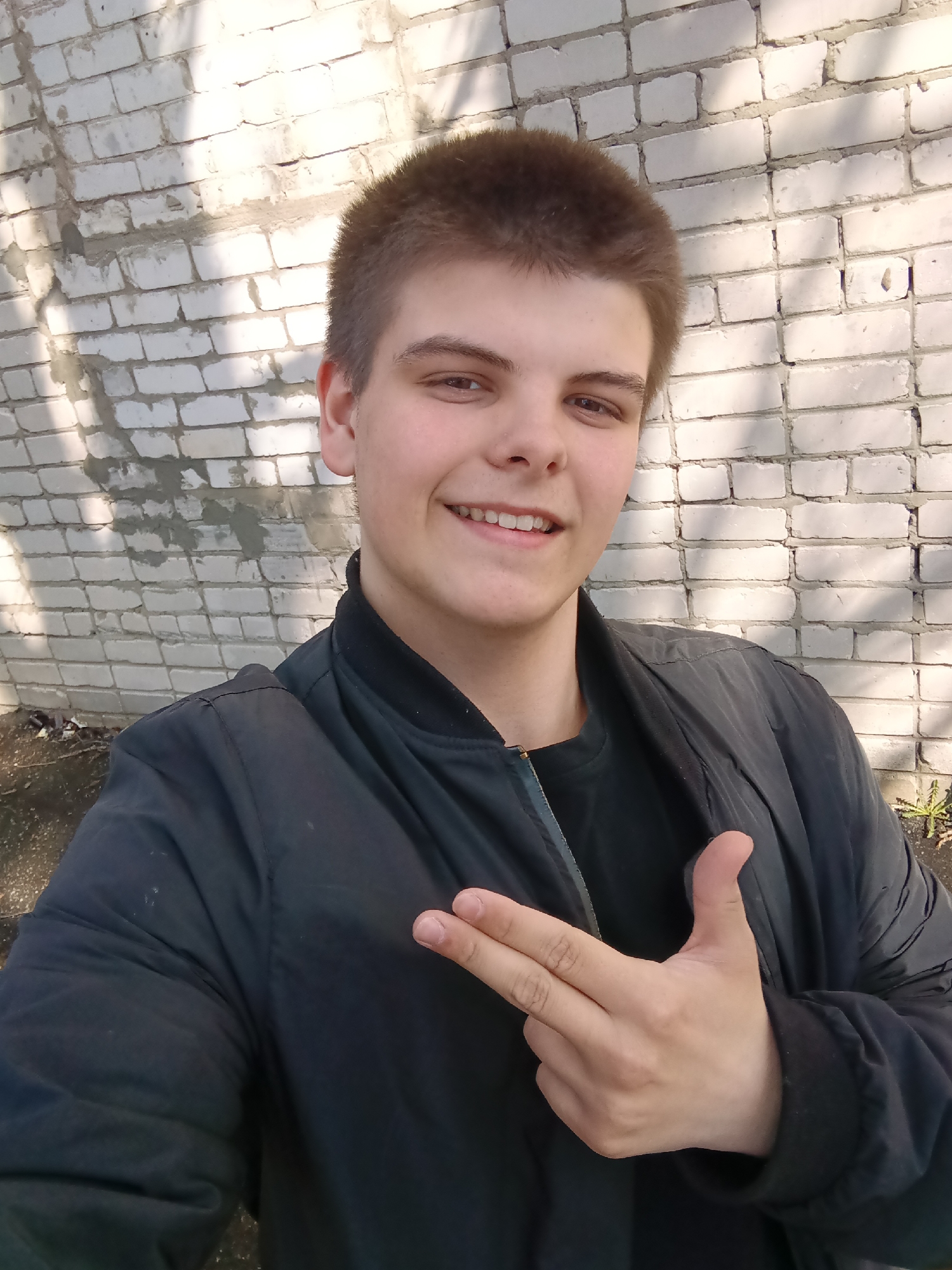 A young man stands in front of a white brick wall, smiling and posing with his hand formed into a gun shape. He wears a dark jacket and a black shirt underneath. The background is a sunlit wall, indicating an outdoor location. The specific location in Красный Маяк, Russia, cannot be determined from the visual data.