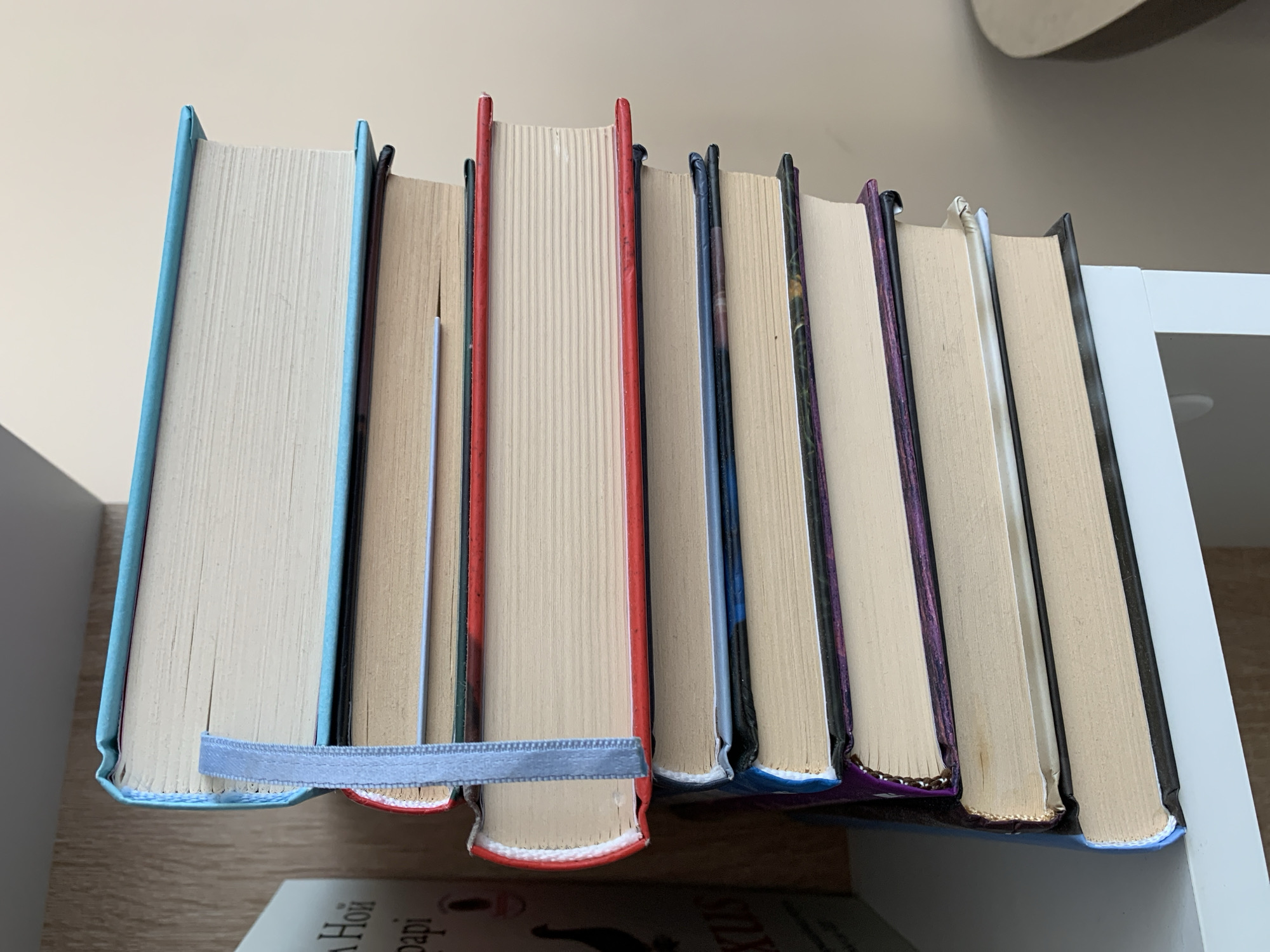 A stack of ten hardcover books is placed on a white shelf, likely in a home library or study. The books vary in color, with spines in shades of blue, red, and purple. A blue ribbon bookmark is visible on one of the top books. The shelf itself is positioned against a light-colored wall, with a wooden surface to the left. There are no people or animals present. The image is cropped to focus on the books, suggesting an interest in their arrangement or content. The scene does not provide any information that could be used to determine its location in Ternopil, Ukraine.