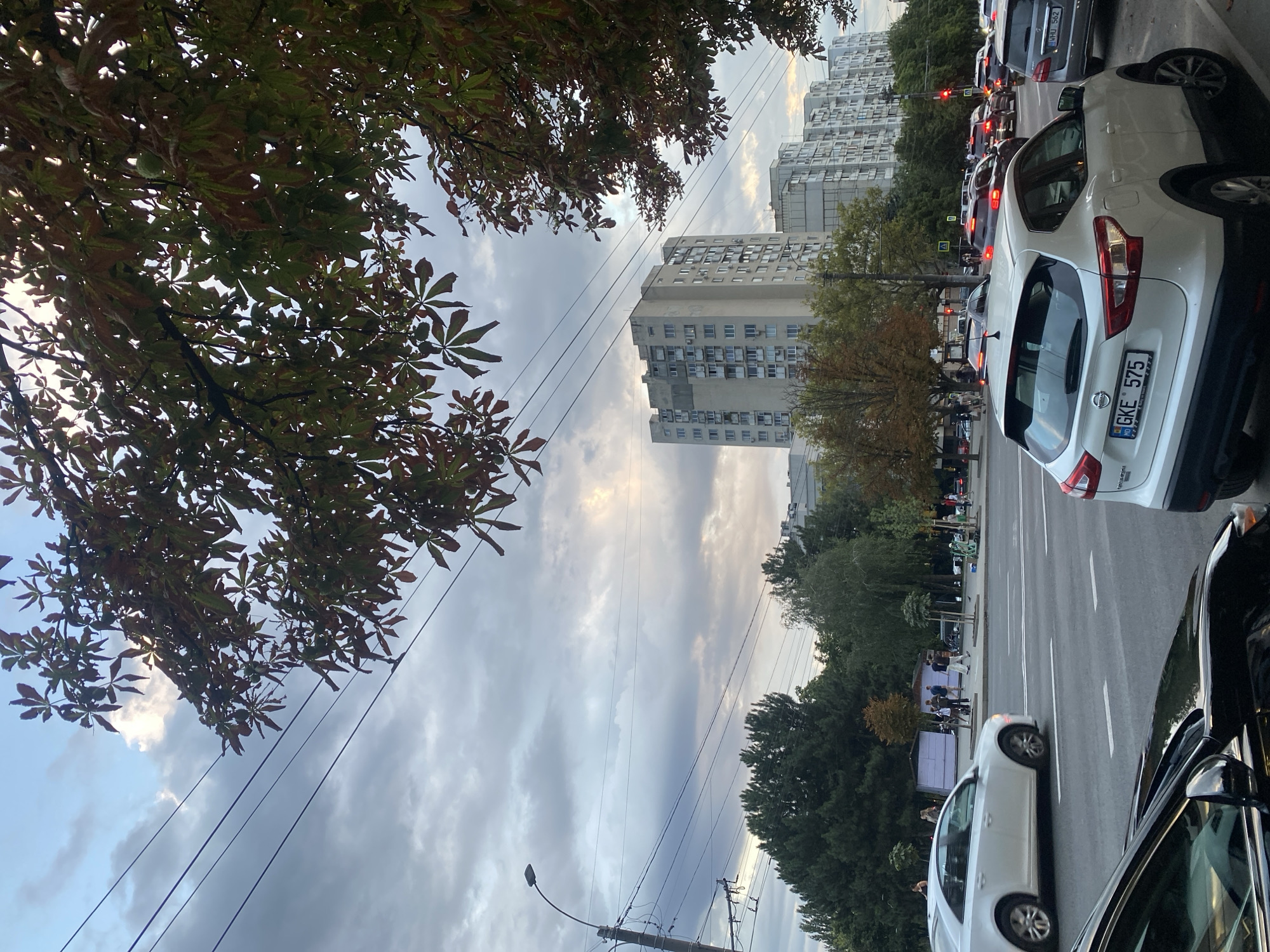 The image shows a street scene in Chișinău, Moldova, with tall apartment buildings in the background. The sky is overcast with some patches of sunlight breaking through. Trees with autumn foliage line the street. Several cars are visible on the road, including a white SUV in the foreground with the license plate GKE 575. People are seen walking on the sidewalk in the middle distance. Power lines crisscross the sky, supported by a lamppost on the left side of the frame. The overall impression is of a typical urban environment during a cloudy day.