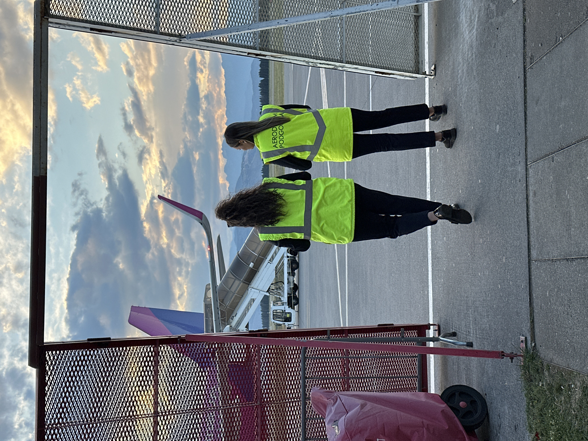 The image shows two people wearing yellow safety vests with "AEROD PODGO" printed on the back. They are standing on an airport tarmac in Donja Cijevna, Montenegro, next to a metal fence and a piece of equipment covered with a red tarp. Beyond them, an airplane with a purple and blue tail fin is connected to a mobile passenger boarding stairs. Mountains are visible in the background under a cloudy sky. The scene appears to be taken from behind the fence, offering a partial view of the tarmac and the aircraft.