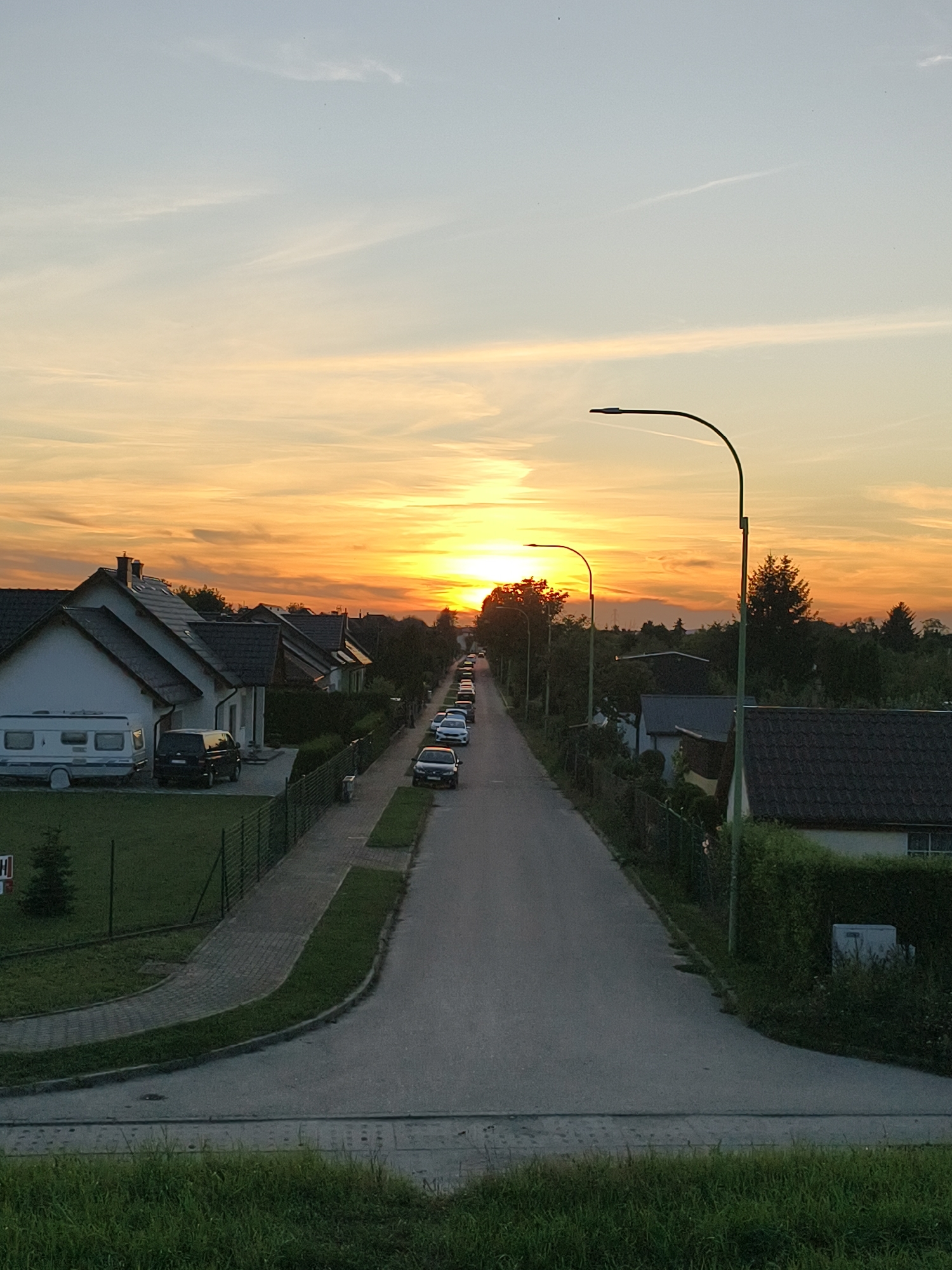 The image shows a suburban street in Tczew, Poland, during sunset. The sky is filled with warm orange and yellow hues, indicating the sun is low on the horizon. The street is lined with houses on both sides, and several cars are parked along the road and in driveways. A white caravan is parked in front of one of the houses. Streetlights are visible along the road. The foreground consists of green grass.