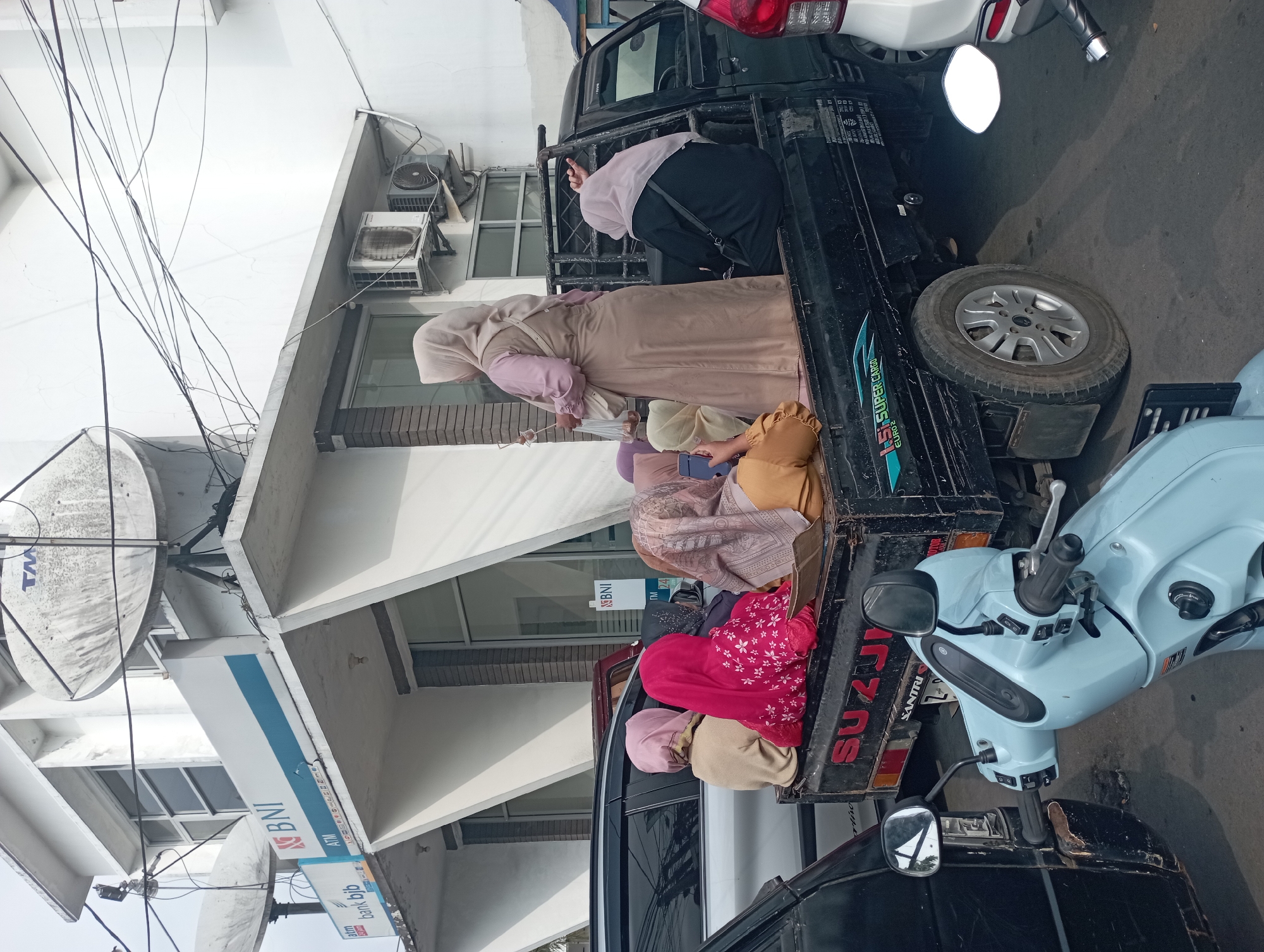 In the city of Tasikmalaya, Indonesia, a group of individuals are gathered outside a building that appears to be a bank, with visible "BNI" and "bank bjb" signage. The foreground is dominated by a light blue scooter, parked next to the back of a black pickup truck. The truck is loaded with several women, all wearing hijabs. One woman, dressed in a light brown abaya and a light pink hijab, stands at the rear of the truck, holding a plastic bag and a mobile phone. Other women are seated in the truck bed, some facing away from the camera, others with their backs to the viewer. The building in the background features large satellite dishes mounted on its roof and air conditioning units on its exterior. The overall scene depicts a moment of everyday life, with people utilizing a pickup truck for transportation.