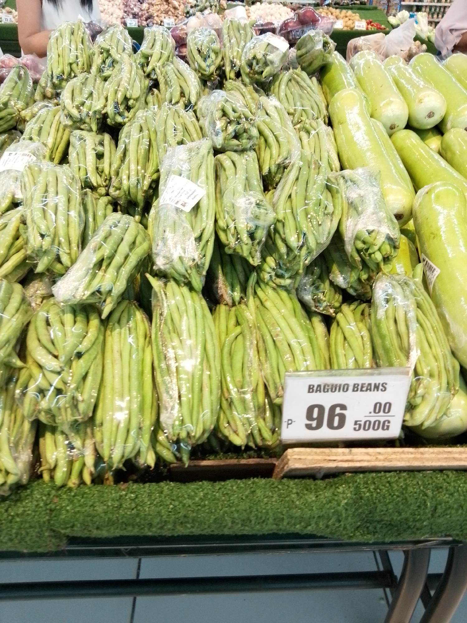 The image shows a display of fresh produce, specifically green beans and some long green gourds, at a market stall. The green beans are packaged in clear plastic and arranged in bundles. A price tag in the foreground indicates "BAGUIO BEANS P- 96 .00 500G". In the background, there are other vegetables and fruits, as well as people browsing. The overall scene suggests a busy marketplace environment. This image was likely taken in a market in Davao City, Philippines, given the presence of Baguio beans, a common produce item in the Philippines, and the general market setup.