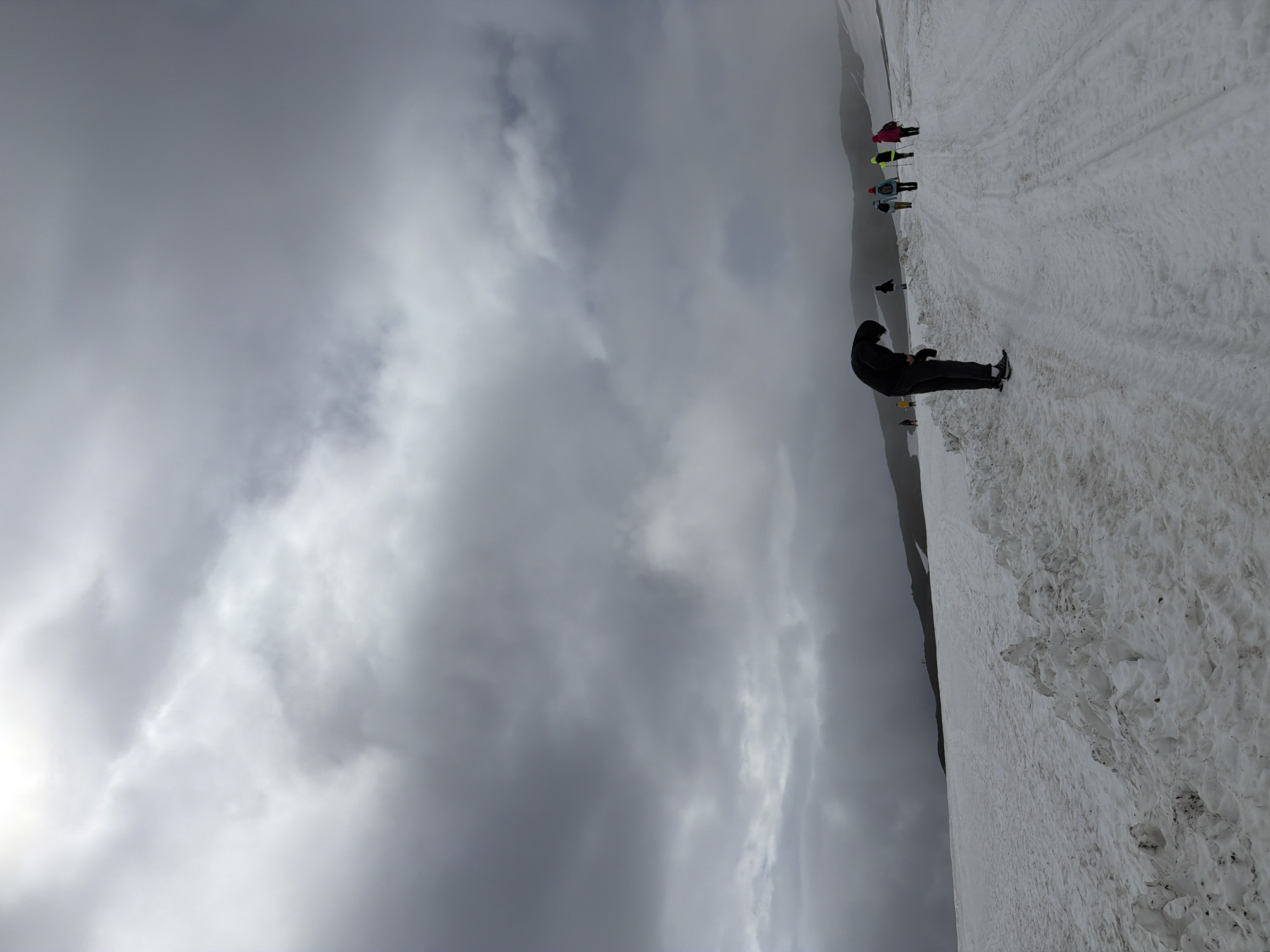 A group of people are walking on a snowy path in a mountainous landscape. The sky is overcast with heavy clouds. The person in the foreground is wearing a black hooded jacket and dark pants, bending over and appearing to examine something on the ground. To their right and further down the path, several other individuals are visible, some wearing colorful clothing and carrying trekking poles. The ground is covered in snow, with tracks visible on the path, indicating recent travel. The background consists of rolling hills and mountains, shrouded in mist and under a dramatic sky. This scene appears to be a winter mountain trek in the region of Nagornyy, Russia, characterized by its cold climate and snowy terrain.