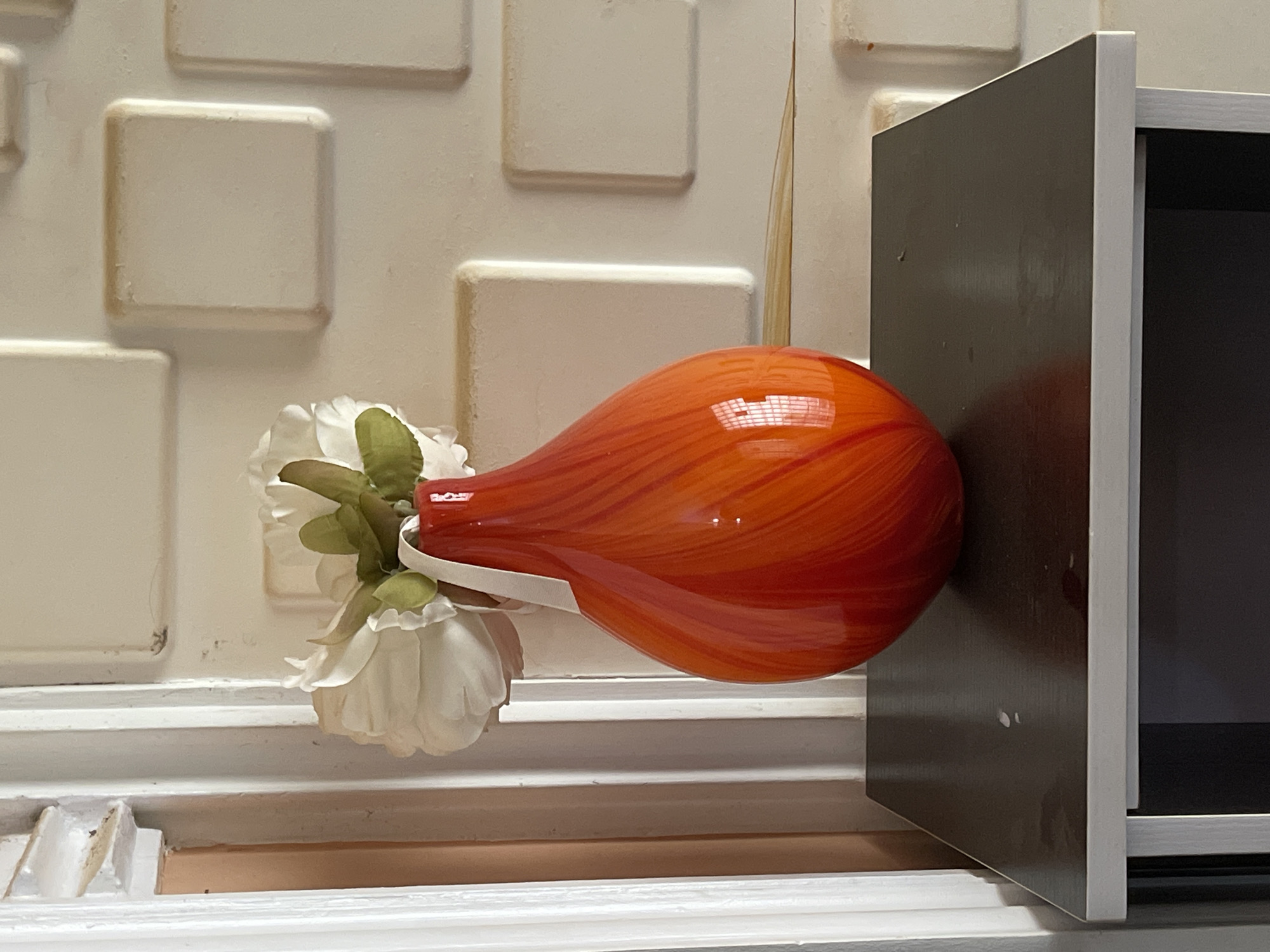 A close-up shot features an orange, tear-shaped glass vase adorned with artificial white flowers. The vase is suspended near a dark brown cabinet door. The background consists of a textured white wall with square-shaped protrusions and a white windowsill with ridges. A thin, straw-like object extends vertically behind the vase. The location within Reifild-Ist, Nigeria, is not determinable based on the image.