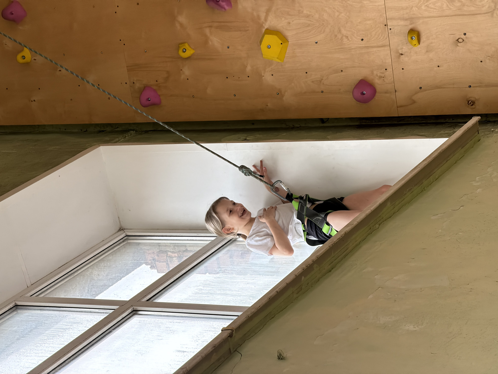 A young child is suspended from a climbing wall in Izhevsk, Russia. The wall is made of plywood with irregularly shaped, colored climbing holds. The child is wearing a harness and is connected to a rope. A large window is visible in the background.
