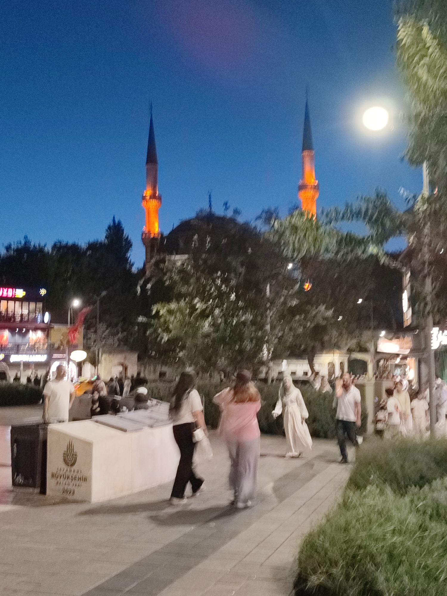 In the city of Üsküdar, Turkey, at dusk, two minarets of a mosque are illuminated with orange light. The sky is a deep blue, fading to lighter hues near the horizon. In the foreground, several individuals are walking on a paved walkway. One woman in a white shirt and black pants walks from left to right. A man in a white t-shirt and dark pants is walking away from the camera, towards the right side of the frame. A person in a long, light-colored garment appears to be walking in the center of the frame. Behind them, a group of people are gathered around a low, white structure with text and a logo visible, identifying it as related to the Istanbul Metropolitan Municipality and the year 2023. Trees and foliage line the right side of the walkway, and a bright light fixture is visible in the upper right corner. Buildings with illuminated windows are present in the background on the left.
