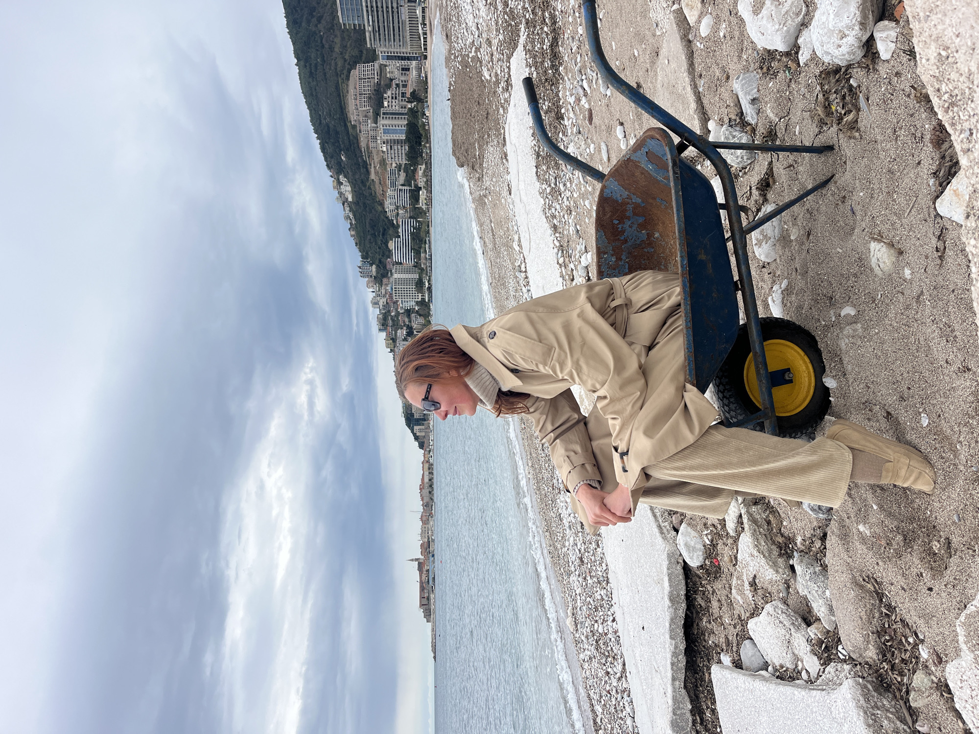 A woman wearing a beige trench coat and corduroy trousers sits on the edge of a blue wheelbarrow near the coast. The location is Budva, Montenegro, suggested by the mountainous terrain and city buildings in the background. The ground is sandy and rocky, near the water's edge, with the sea and cloudy sky visible in the background. The woman is wearing sunglasses. The wheelbarrow has a yellow wheel and rusted interior.