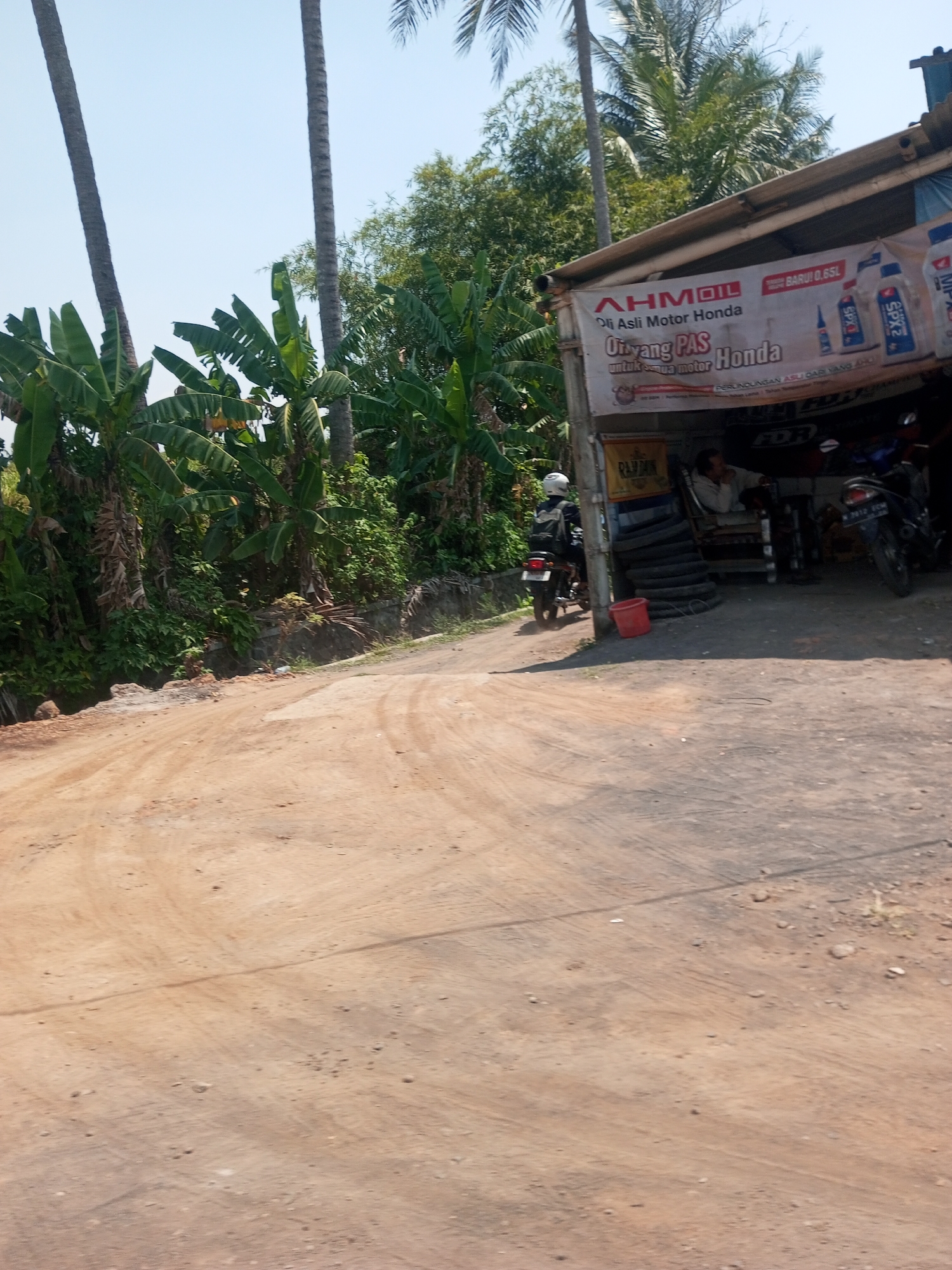 The image captures a roadside scene in Singosari, Indonesia, on a clear day. In the foreground, a dirt road shows tire tracks and scattered pebbles. To the left, lush green banana trees and tall palm trees line the road. In the background, a modest shop with a corrugated metal roof displays a banner advertising "AHMOIL Oli Asli Motor Honda" and other motorcycle-related products. Two motorcycles are visible, one parked outside the shop and another being ridden by a person wearing a white helmet and a backpack, heading away from the camera. A man is seated inside the shop, leaning back in a chair. A red bucket sits near a stack of tires. The overall impression is one of a rural or semi-rural commercial establishment.