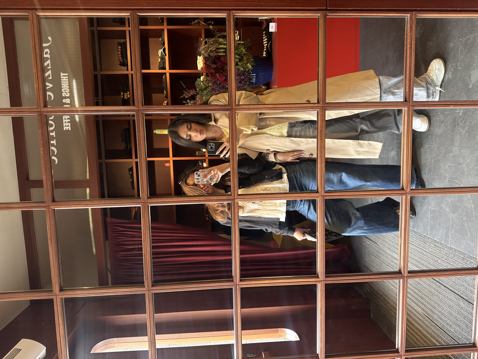 Two women are reflected in a multi-paneled mirror, taking a selfie. One woman wears a trench coat and jeans, while the other wears a black jacket and jeans. Shelves with merchandise and decorative plants are visible in the background, along with a red curtain and a sign with text. The environment suggests a retail or commercial setting, possibly in Yerevan, Armenia.