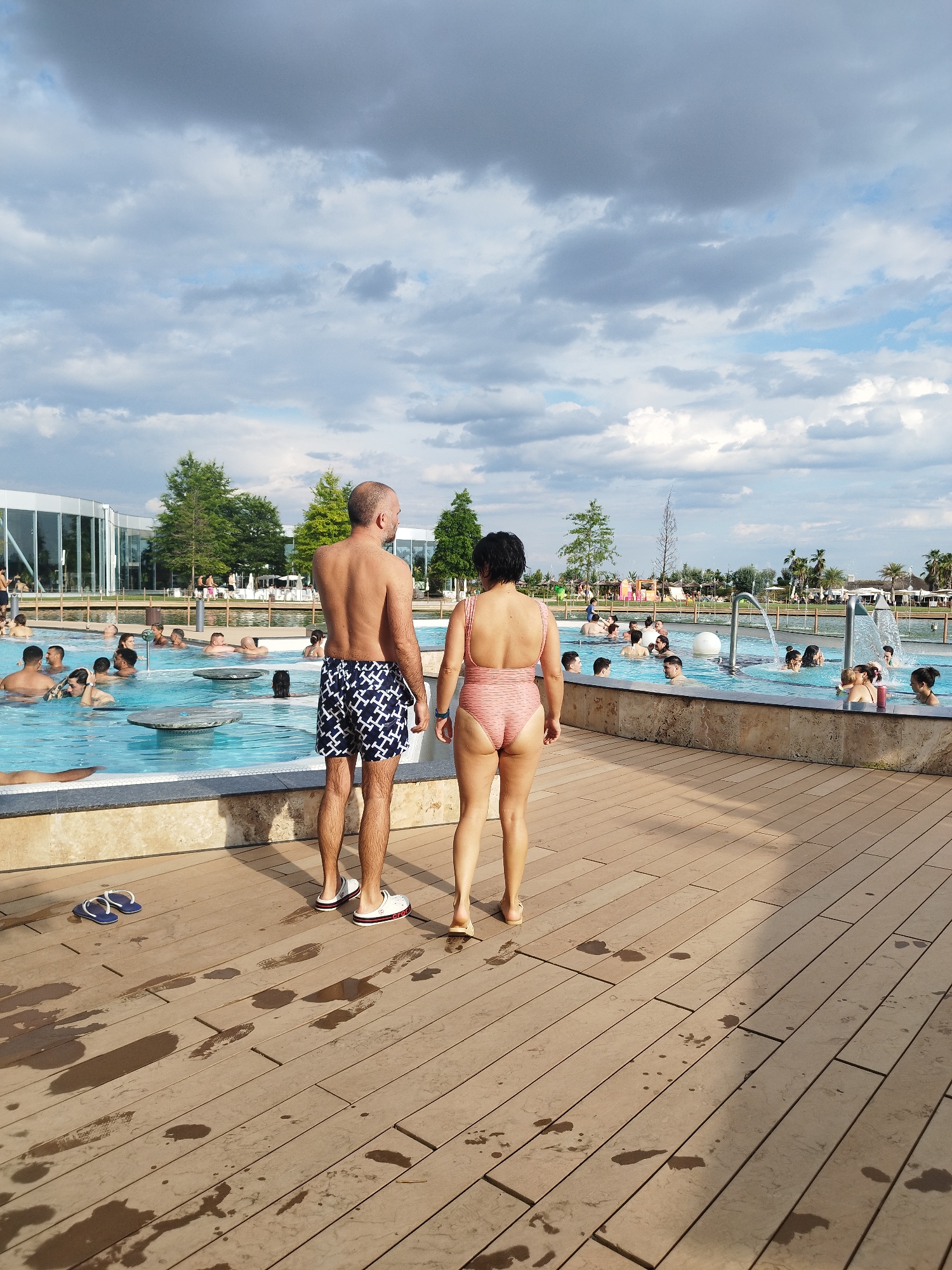 This media file shows an outdoor swimming pool area on a partly cloudy day. The sun is casting shadows, suggesting it is likely afternoon. The scene is populated with many people of various ages swimming, relaxing in the water, and socializing.
In the foreground, a man and a woman are standing on a wooden deck, facing away from the camera and towards the pool. The man is wearing patterned swim trunks, and the woman is wearing a pink, striped one-piece swimsuit. They appear to be observing the activity in the pool. The wooden deck is wet in several places, with visible footprints. A pair of blue flip-flops lies on the deck near the man's feet.
The pool itself is large and features several integrated circular tables or resting areas in the water. In the background, there is a modern building with large glass windows, possibly a spa or leisure facility. Green trees and palm trees are visible around the perimeter of the pool area. There are also some children's play structures visible in the distance.
The overall mood seems relaxed and leisurely, typical of a day at a water park or resort. The presence of umbrellas and lounge chairs in the background suggests a place designed for recreation and relaxation. The location context provided is Săftica, Romania. No visible text is present in the image.