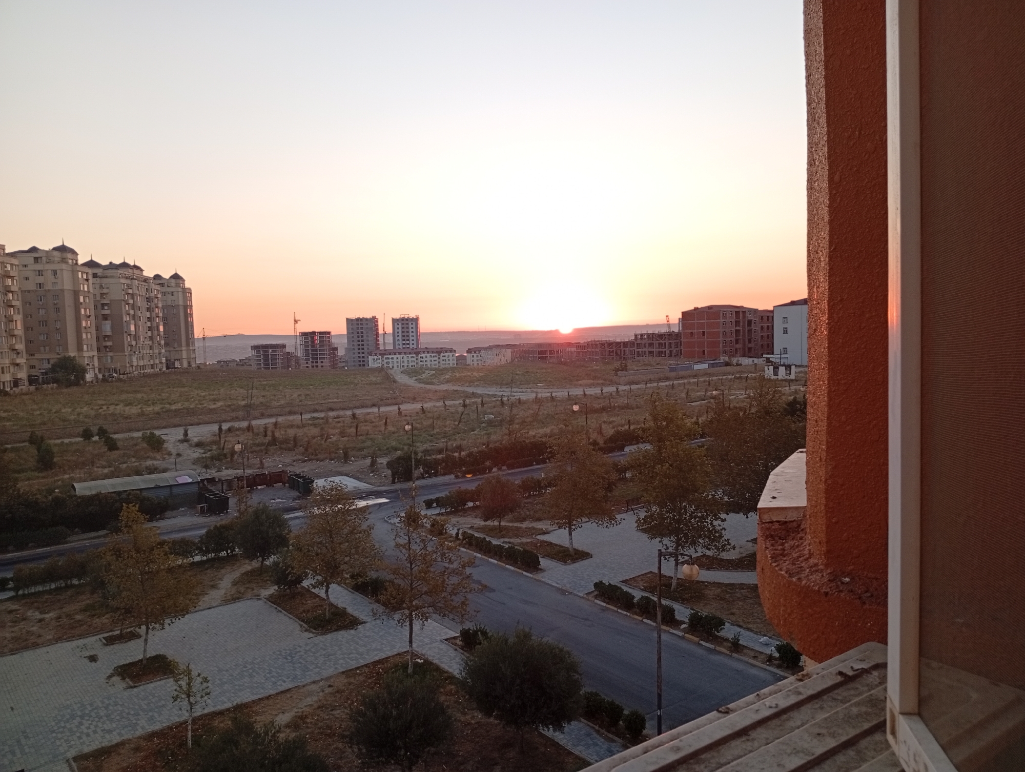 This is a medium-shot, eye-level photograph taken during sunset, capturing a view of the city of Masazir, Azerbaijan, from a balcony. The foreground shows the edge of the balcony with a textured orange railing and a window frame. In the middle ground, a street runs through a landscaped area with trees and pedestrian walkways paved with gray interlocking bricks. The street itself is paved with asphalt. Beyond the street, there are more trees and patches of dry grass. In the background, several multi-story buildings, some of which appear to be under construction with visible scaffolding, dominate the horizon. The sky is a pale orange and yellow gradient, indicating the sun is low, with the sun itself partially visible near the horizon. The overall impression is of an urban developing area at dusk.