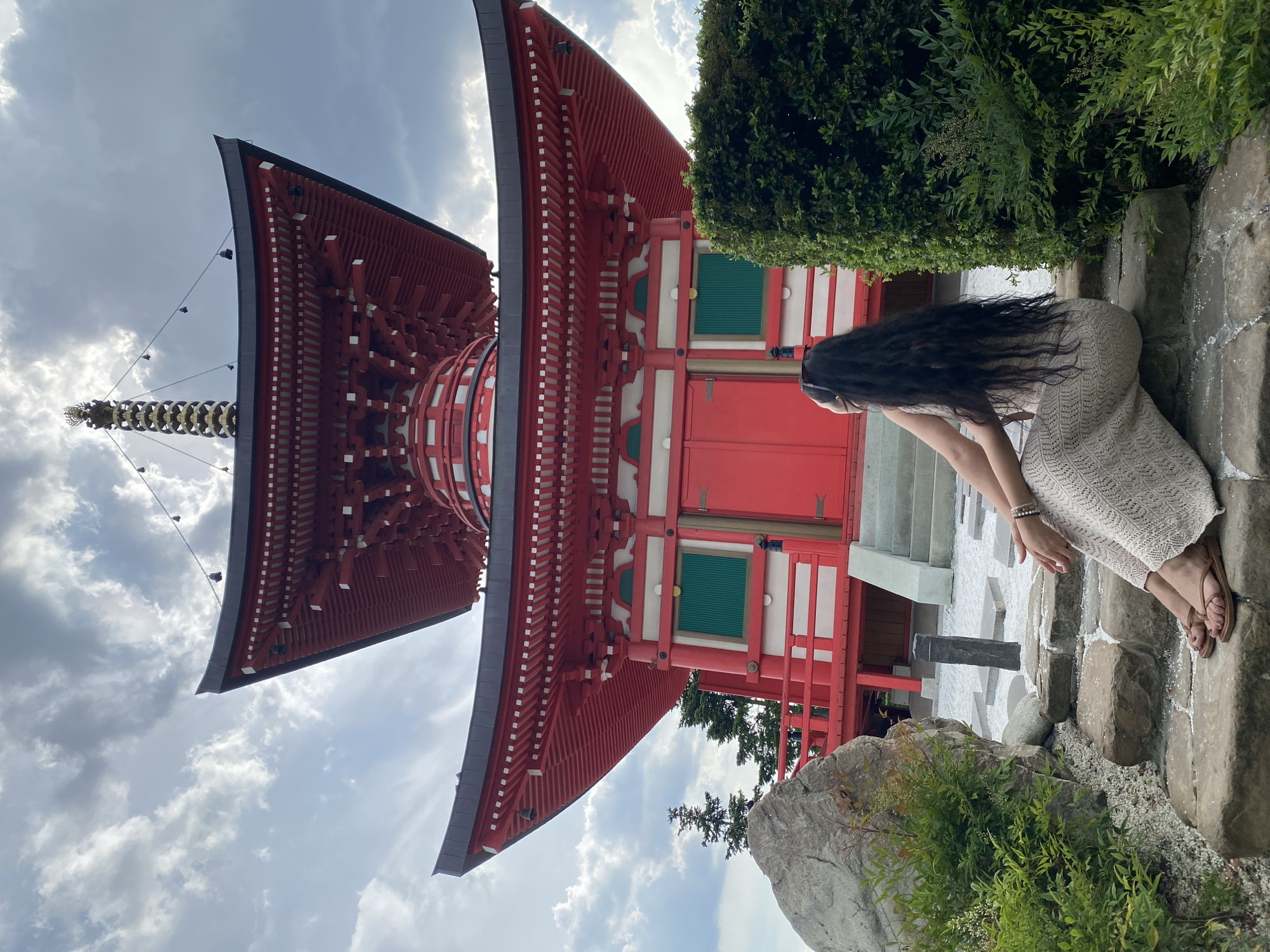 A low-angle, full-shot photograph depicts a female individual seated in the foreground, observing a multi-tiered red pagoda in Krasnodar Park, Krasnodar, Russia. The pagoda, a replica of a Japanese architectural style, features two prominent red roofs with black eaves and white detailing, topped by a golden finial (sōrin). The lower section of the structure is characterized by large red doors, white walls, and two recessed green-paneled window or door sections. The sky above is overcast with scattered grey clouds. The female subject, positioned with her back to the camera, is seated on rough-hewn stone steps. She has long, dark, wavy hair and is wearing a light-colored, patterned maxi dress and brown open-toe sandals. A bracelet is visible on her left wrist. The immediate environment includes a white pebble garden interspersed with additional stone elements, a large grey boulder, and various green shrubs and bushes. A manicured green hedge partially obscures the right side of the foreground. Evergreen trees are visible on the left side, behind the pagoda's base.