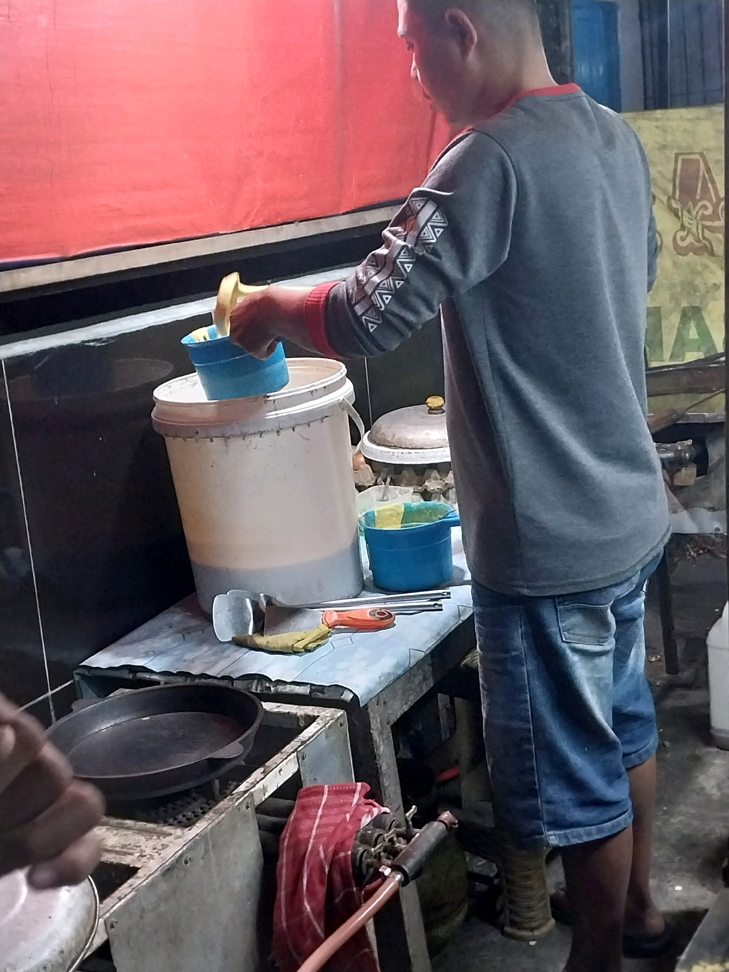 A man in a gray sweatshirt and blue jeans is standing behind a table with various objects on it. He is holding a blue plastic scoop over a large white bucket with a gray bottom, appearing to be transferring something into it. The table also holds a smaller blue bucket with yellow contents, a metal spatula, a yellow glove, and an orange handled tool. Behind the table, a large metal pan is visible on a stove-like structure. To the right of the table, a part of a gas cylinder is visible, draped with a red and white striped cloth. The background shows a red tarp covering the upper portion of the scene and a yellow sign with a partially visible logo on the right. The scene appears to be outdoors or in an open-air establishment, possibly a food stall or a workshop, given the cooking equipment and industrial-looking elements. The location is identified as Nglegok, Indonesia.