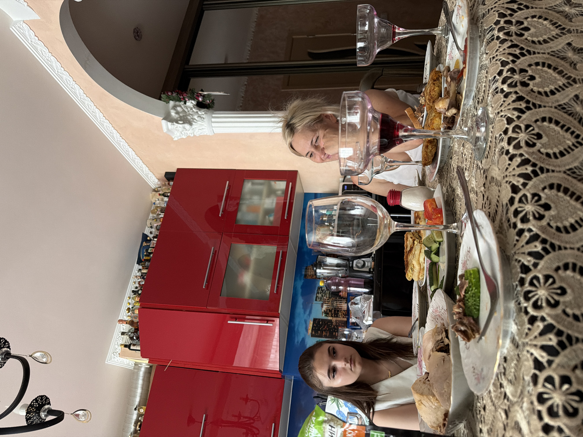 In a home setting in Durlești, Moldova, two women are seated at a table laden with food and drinks. The woman on the left, a young female with dark hair, wears a white top and is looking down with a somber expression. On the right, an older woman with blonde hair is leaning her head on her hand and smiling, appearing engaged in conversation. The table is covered with a beige lace tablecloth and features several plates of food, including what appears to be roasted meat, vegetables, and pastries. Wine glasses and a salt shaker are also visible. Behind the table, red kitchen cabinets are mounted on the wall, and a cityscape mural is depicted. The background also shows a large archway with a decorative column and a door with a mirrored surface. The overall ambiance suggests a casual meal or gathering within a residential interior.