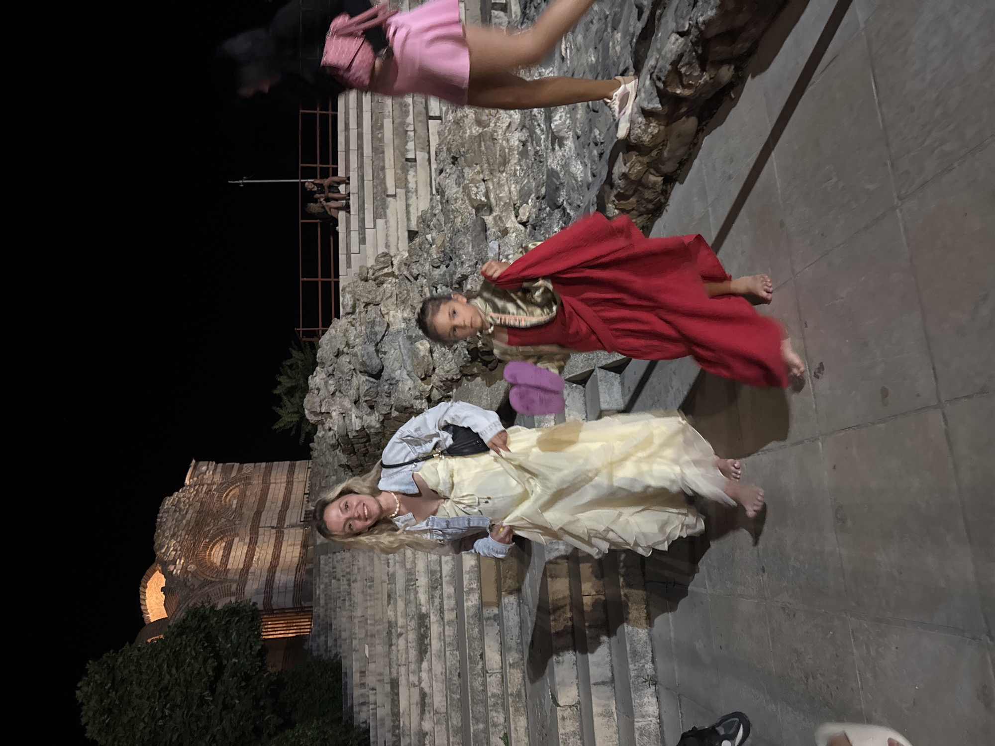 At night in Nesebar, Bulgaria, three people are visible. On the left, a woman with blonde hair wears a pale yellow ruffled dress and a denim jacket, holding her dress and a black purse. To her right, a young girl in a red dress and gold vest stands barefoot, holding a red cloth. Further right, a person in a pink skirt and black top walks away from the camera. Behind them, stone steps and a ruined brick structure are illuminated. Two other people are seated on steps in the background.