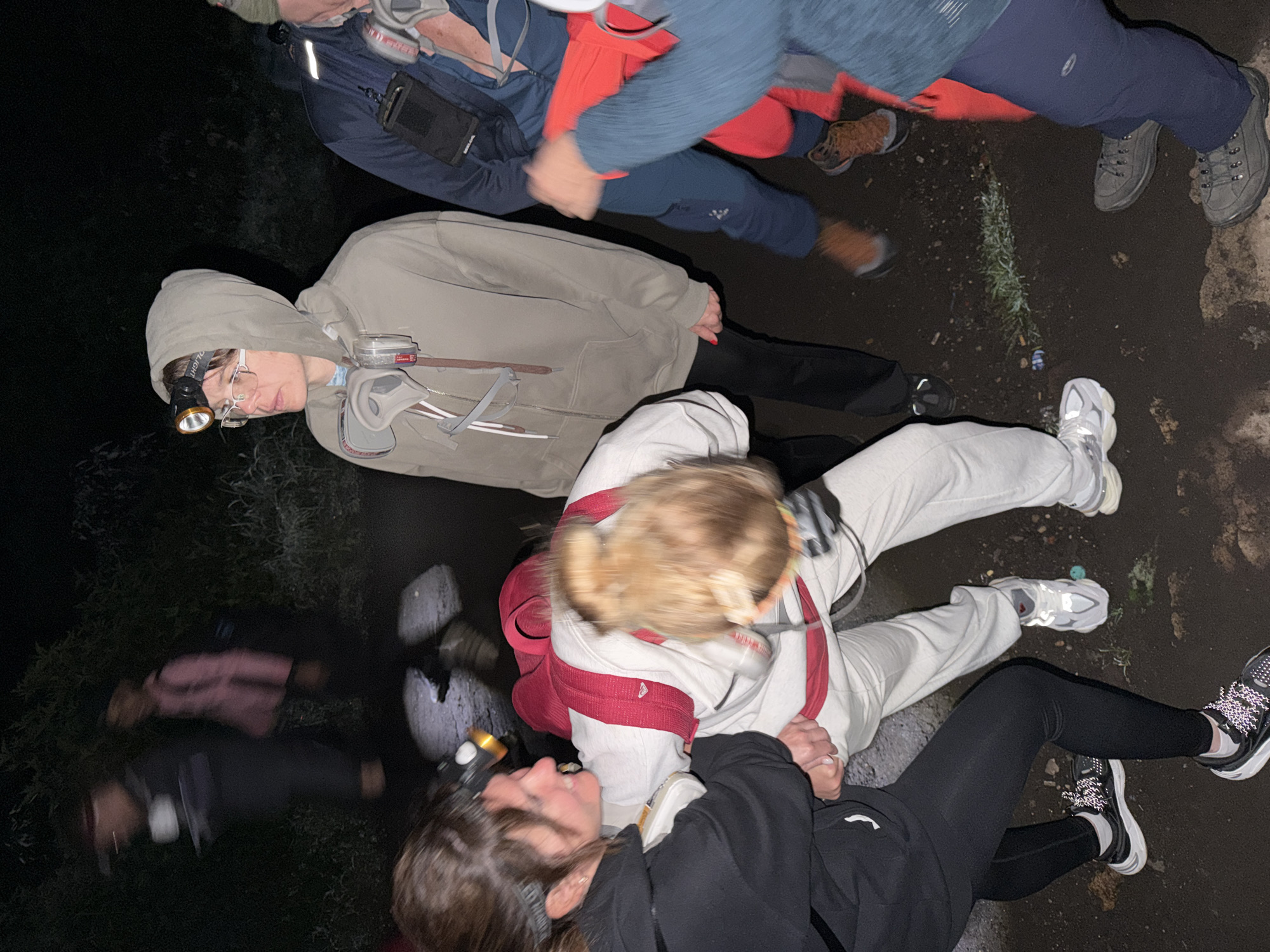 A group of people are gathered together in an outdoor setting, likely at night, as evidenced by the darkness and the use of headlamps. They appear to be on some type of ground surface that looks like a trail or path. The people are dressed in casual clothing including hoodies, pants, and sneakers. Some of the individuals are wearing backpacks and headlamps. The faces of the people are partially visible but not easily identifiable due to the darkness. Some interaction is occurring within the group, as some people appear to be holding hands. The precise location is not discernible beyond being outdoors, and the city/country context provided is Unknown, Indonesia.