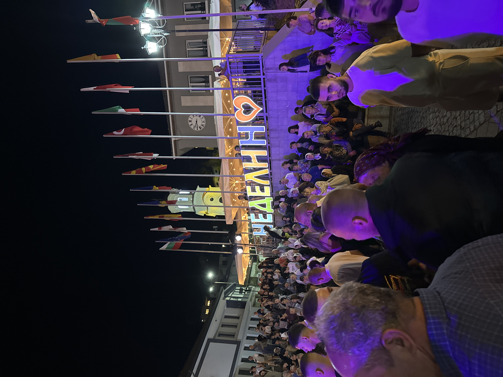 The image depicts an outdoor nighttime event in the city of Nedelino, Bulgaria. In the background, a large illuminated sign with the word "НЕДЕЛИНО" and a heart symbol is prominent, suggesting a festive occasion. A crowd of people is gathered, facing towards the sign and the area beyond, possibly for a performance or a public gathering. Several national flags are displayed on poles in the background, adding to the celebratory atmosphere. A clock tower is visible on a building behind the flags, indicating the presence of significant architecture within the city. The foreground shows the backs of several individuals, indicating the perspective of someone within the crowd. The lighting suggests artificial illumination, with spotlights and the illuminated sign contributing to the ambiance. The overall scene conveys a sense of community and public engagement in Nedelino.