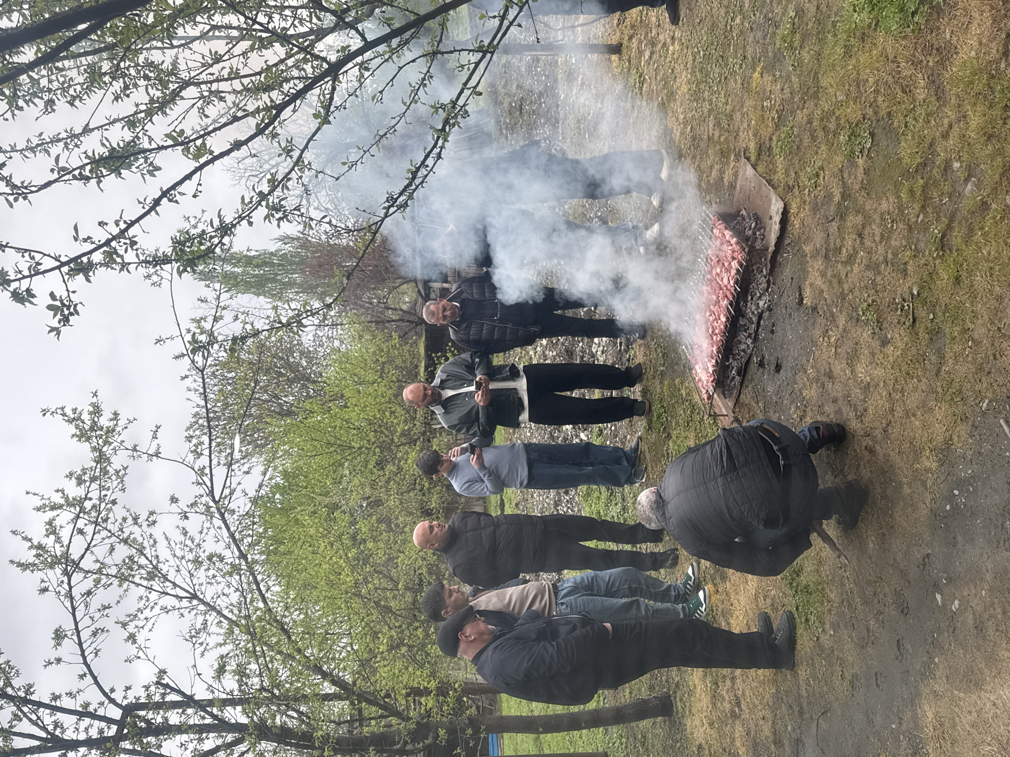 The image depicts a group of approximately seven men gathered outdoors around a cooking fire in Kartubani, Georgia. Meat skewers are laid over a crude fire pit. Smoke obscures the background. The men are of various ages, and all are dressed in casual clothing. One man is kneeling by the fire. The surrounding environment consists of grass, trees, and overcast skies. A wooden structure is in the far left corner of the image.