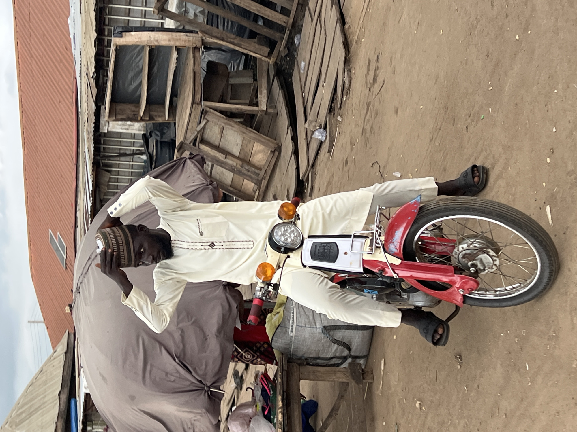 The image shows a man sitting on a red and white motorcycle in what appears to be an outdoor market in Oko Amakom, Nigeria. He is wearing a cream-colored traditional outfit and a striped cap, which he is adjusting. The motorcycle has chrome handlebars, orange turn signals, and a black seat. In the background, there are market stalls with tarpaulins, wooden structures, and various goods. The ground is dirt. The sky is overcast.