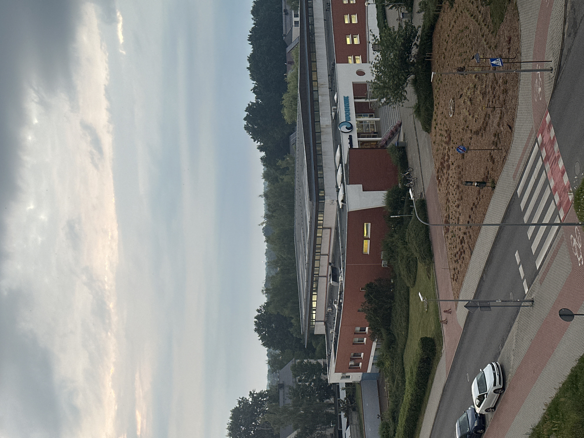 This is a high-angle, medium shot of a building in Żary, Poland, on an overcast day. The building, identified by the "WODNIK" logo, appears to be a public facility, possibly a sports center or aquatic facility, given the logo's wave-like design. The building has a modern architectural style with red brick accents and a white facade. There are cars parked along the street in front of the building, and a pedestrian crossing is visible. The surrounding area is green, with trees and landscaping. The sky is cloudy, and the lighting suggests it is either early morning or late evening.