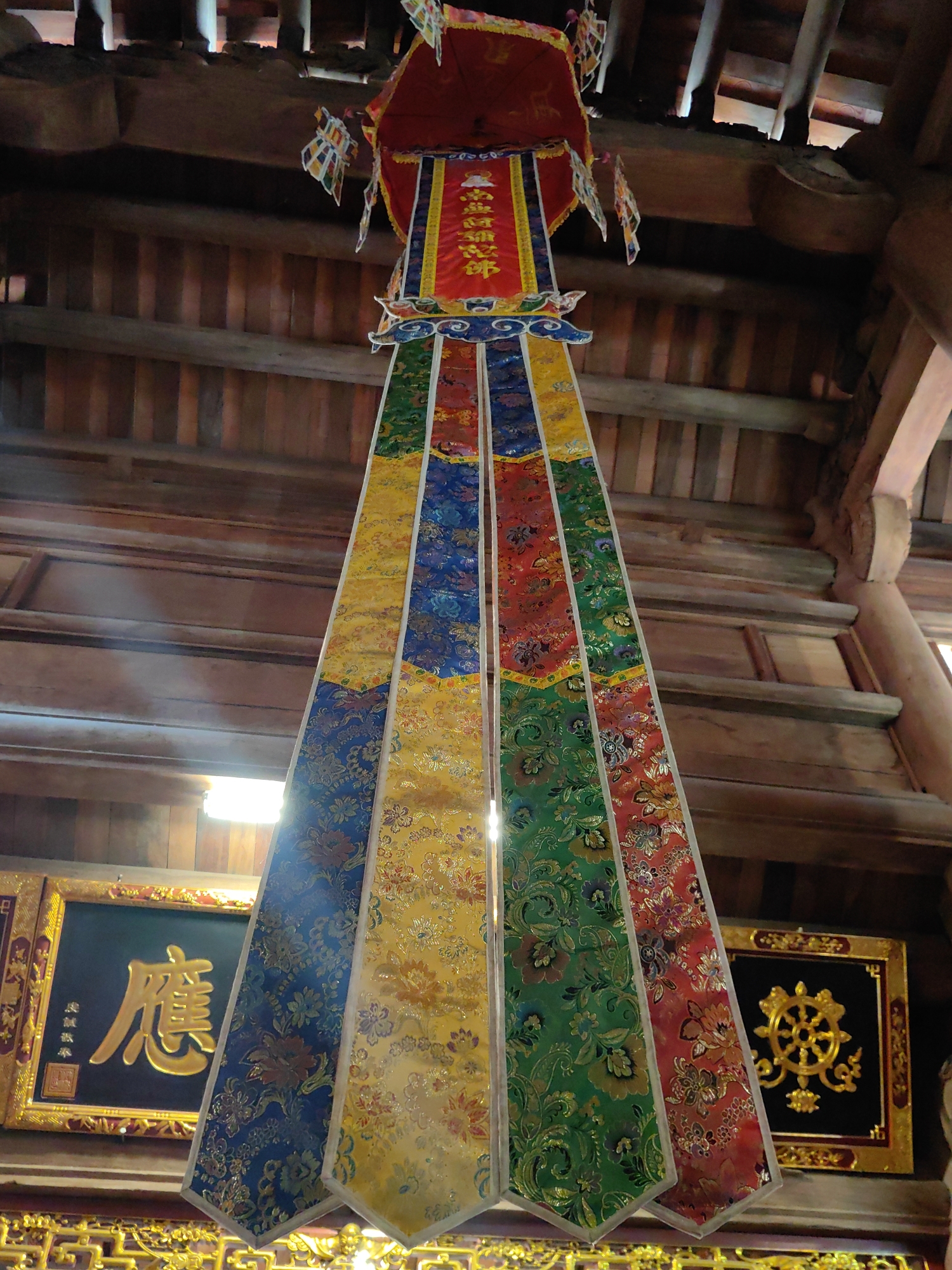 A low-angle, full shot features a multicolored fabric decoration hanging from a wooden ceiling. The fabric is divided into four vertical sections of blue, yellow, green, and red. Each section features an ornate floral pattern. Above the fabric is a cylindrical red, yellow, and white topper. Small paper flags hang off the top of the topper. In the bottom left is a black square framed with gold, which is adorned with gold lettering in Chinese. To the right of the fabric, in the bottom right corner, is another similar black square framed with gold, which is decorated with a golden circular symbol. The wooden ceiling of the structure is visible in the background. The photo was taken in the city of Hong Gai Ward, Vietnam.