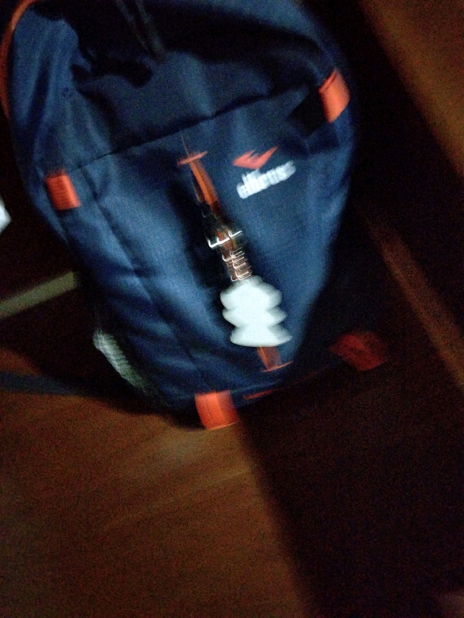 The image shows a dark blue backpack with orange accents. A zipper pull, which appears to be a small, white, multi-tiered ornament, is attached to a zipper on the backpack. The backpack is lying on a wooden surface. The lighting in the image is dim, with a blur effect suggesting movement. The overall scene is not specific to any particular location in the city of Jenangan, Indonesia.