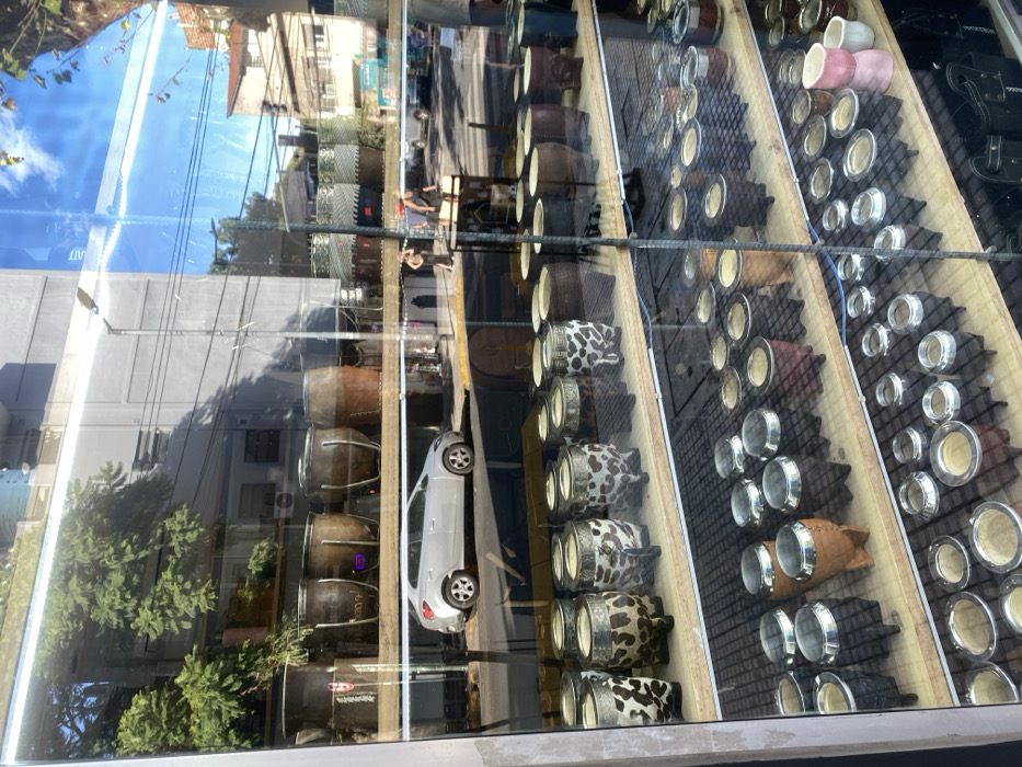 A shop window displays rows of mate cups, featuring various designs from plain silver to animal prints. Reflections in the glass reveal a street scene with buildings, trees, and a parked car. Sunlight glints off the cups, highlighting the display against the backdrop of the urban environment.