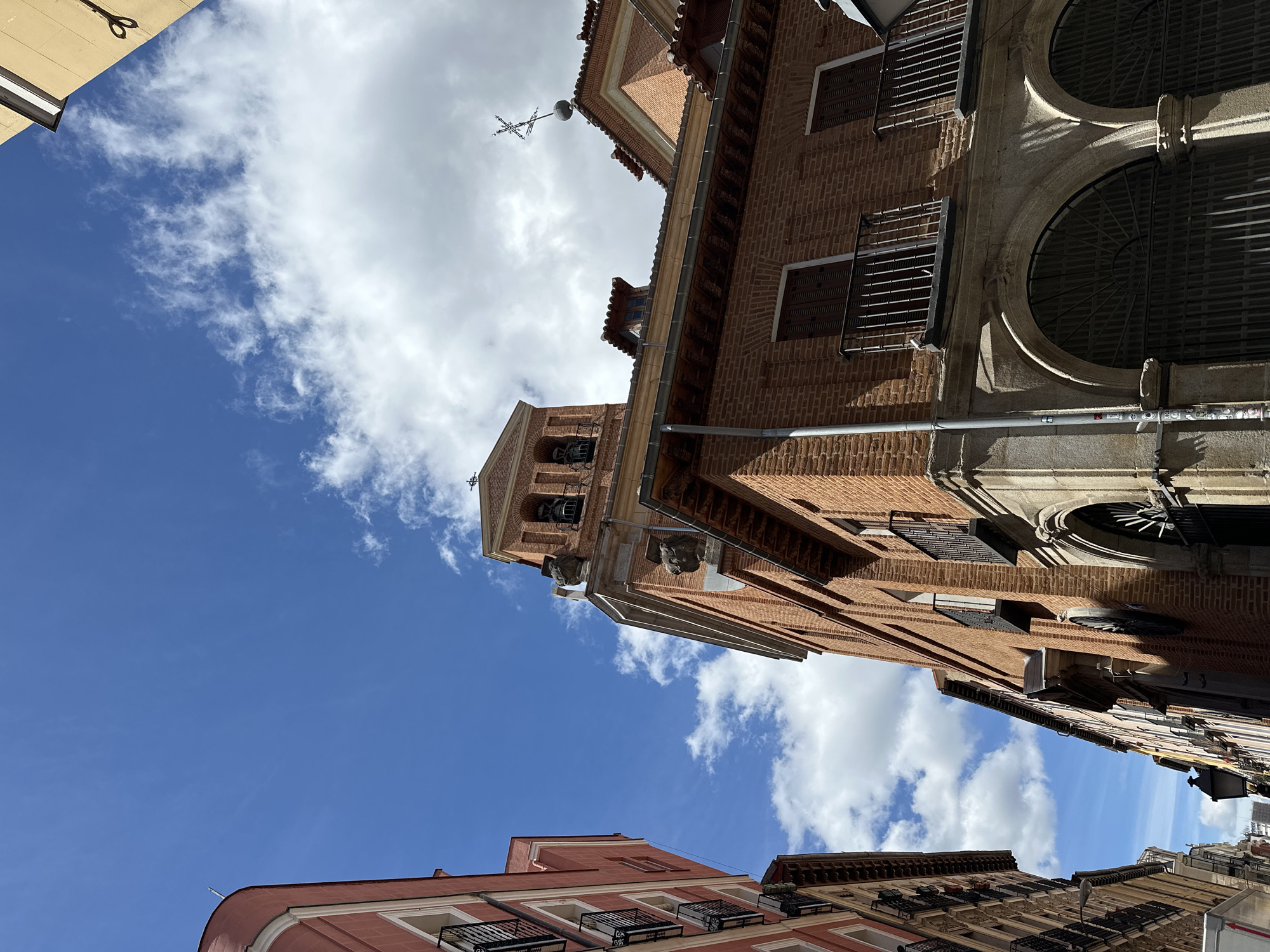 The image depicts a street in Madrid, Spain, as seen from a low angle, looking upwards. The sky is visible, with a mix of blue sky and white, puffy clouds. Buildings line the street. The main building is made of red brick with a tower and arched windows with decorative metal grilles. Adjacent buildings show variations in architecture, including one with a reddish facade and balconies. The image provides a sense of the narrow streetscapes typical of older parts of Madrid.