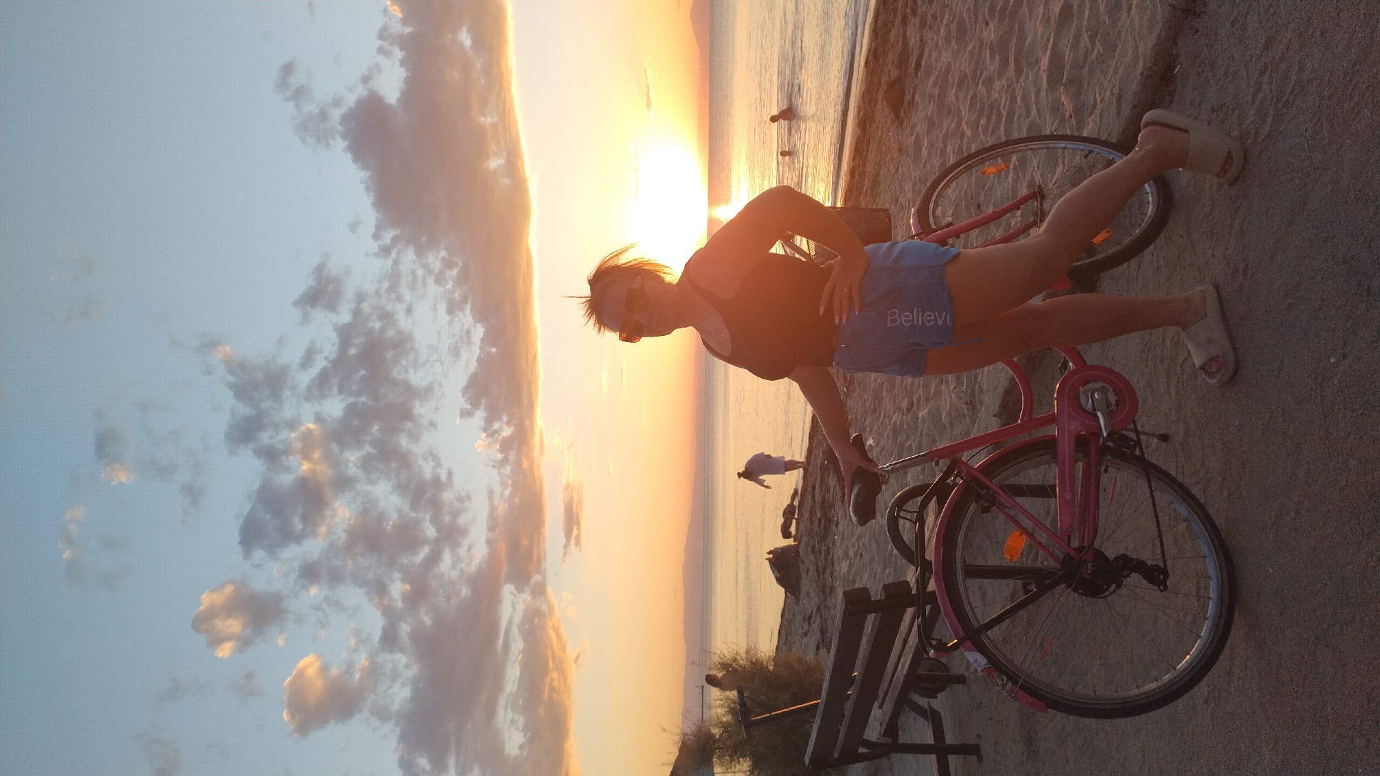 An outdoor, full shot captures a woman standing on a sandy beach in Perea, Greece, with a bicycle beside her, facing right. The woman, wearing a black tank top and blue shorts, stands with her left hand on her hip and her right hand resting on the bicycle's handlebars. She is looking to her right, towards the sun setting over the sea. The sea is calm, and a few distant figures are visible wading in the water. The sky is a gradient of blue and orange, with clouds scattered across it. A wooden bench is visible to the left of the frame, and beyond that, some shrubs. The overall atmosphere is serene and picturesque, indicative of a sunset on the coast.