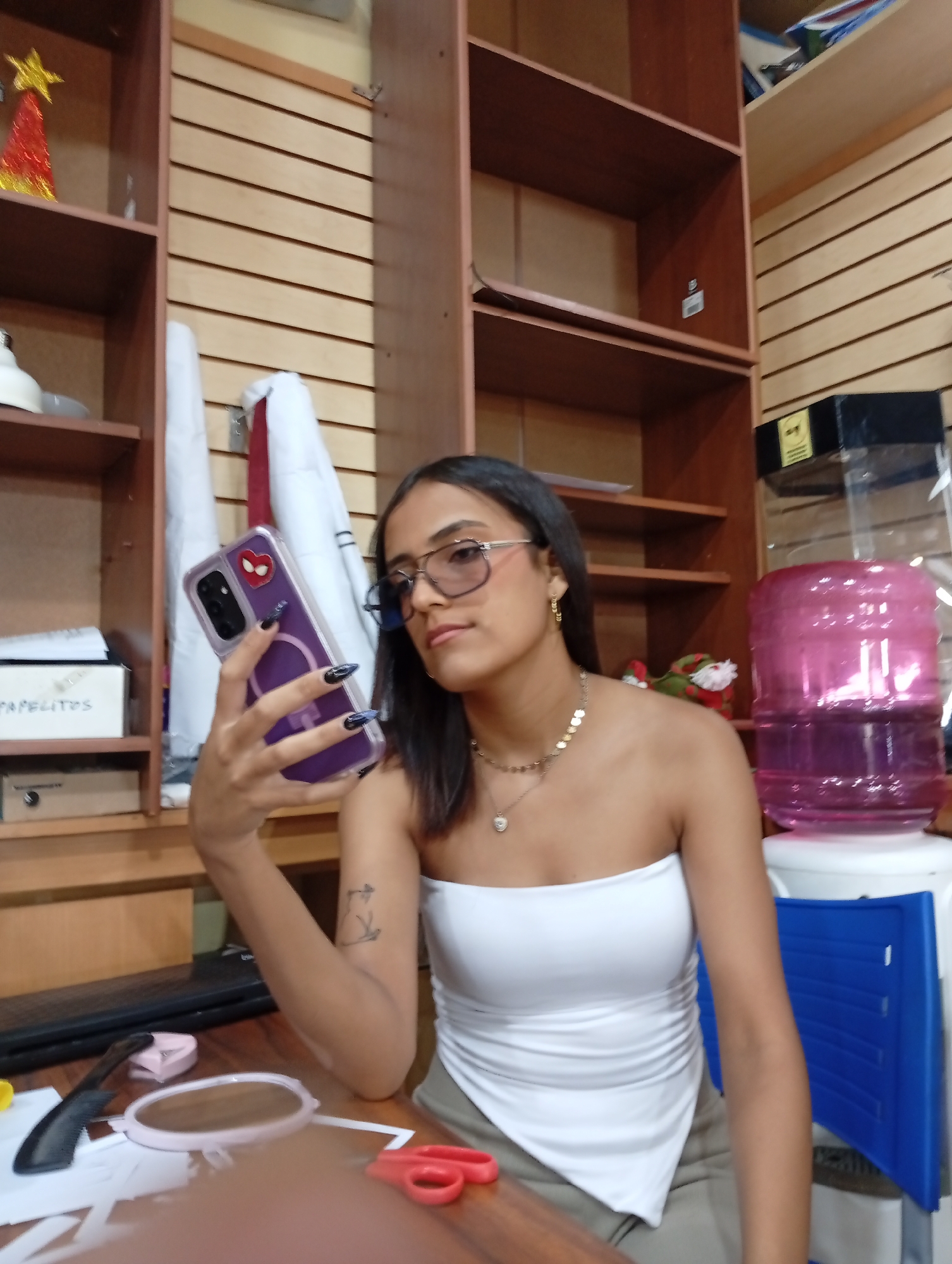 A young woman is taking a selfie in a room with shelves. She is wearing a white tube top, glasses, and jewelry. She holds a purple phone with a Spiderman sticker on it. The room has wooden shelves with various items, including a Christmas decoration, boxes, and a water dispenser with a pink bottle. A blue chair is also visible. The location is identified as Juan Griego, Venezuela. The scene appears to be indoors, with items scattered on a table. A comb, mirror, and scissors are on the table. The subject is actively taking a selfie.