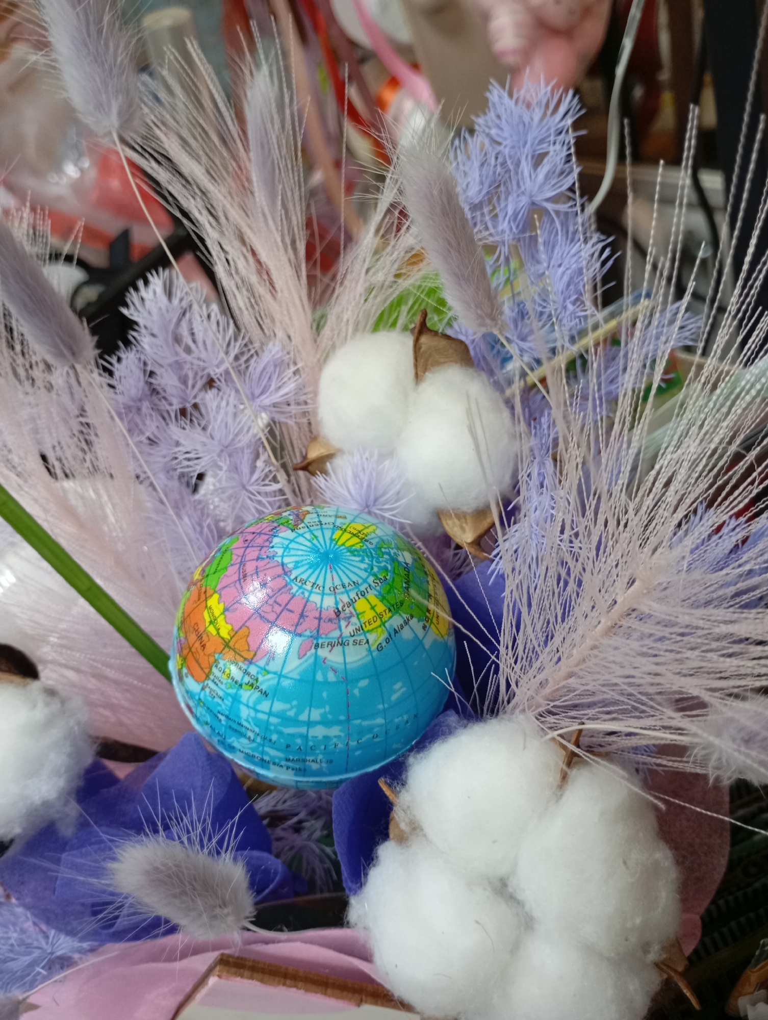 The image shows a small decorative arrangement. A toy globe with visible regions like "RUSSIA," "CHINA," "CANADA," and the "ARCTIC OCEAN" is centrally placed. Around the globe, there are fluffy cotton boll decorations and feathery, possibly artificial, plant accents in white and lavender colors. Purple tissue paper is also visible.