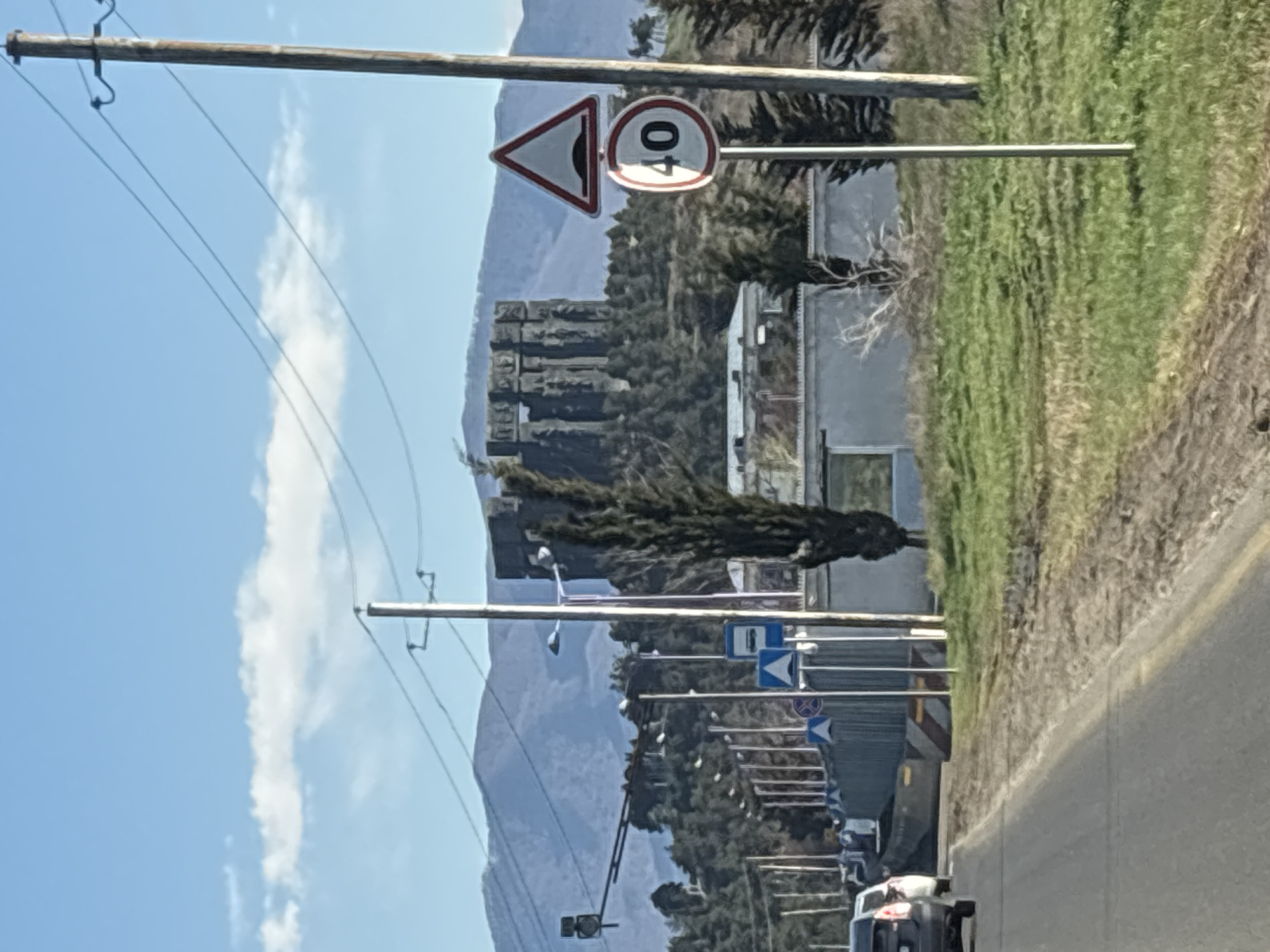 The image depicts a roadside scene on the outskirts of Tbilisi, Georgia. The view is from a vehicle traveling on a paved road. Roadside infrastructure, including utility poles with power lines and numerous road signs, is visible. A triangular warning sign depicting a bump in the road, as well as a circular speed limit sign indicating 40 km/h, are prominent. In the background, a rugged mountain range is partially obscured by scattered trees and vegetation. A structure that resembles a historical fortress or watchtower stands on the mountainside. There are cars on the road. The sky is blue with scattered clouds. The scene suggests a rural or suburban setting, with a blend of infrastructure and natural landscape.