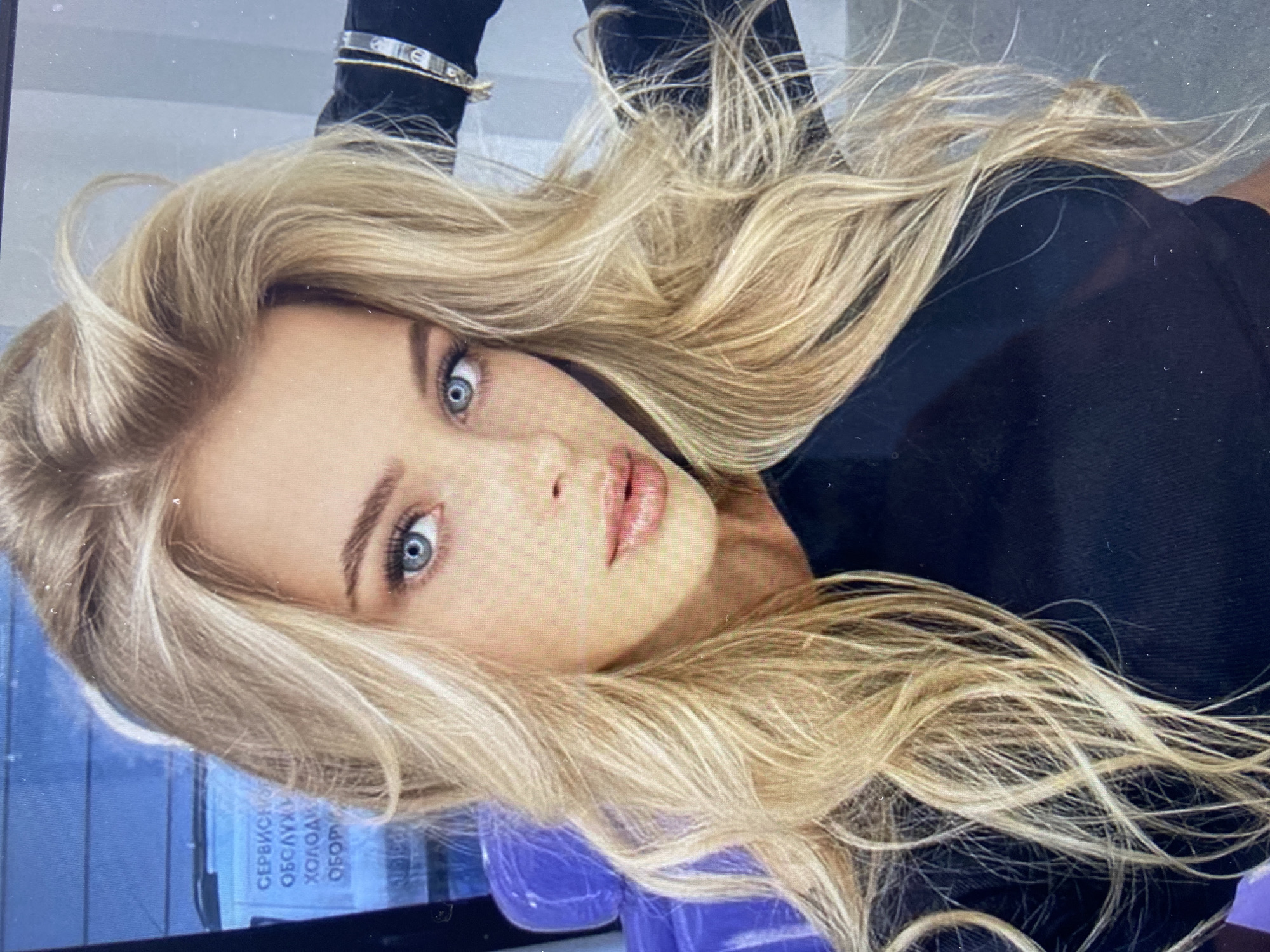 A close-up, medium shot captures a young woman with blonde, wavy hair and striking blue eyes. She is wearing a black top and has a bracelet on her right wrist. Her hair is styled loosely around her face, and she appears to be looking directly at the camera. In the background, a blue, cushioned seat is visible to the left, and text on a blue surface suggests an indoor setting. The lighting is bright and even, highlighting the woman's features.