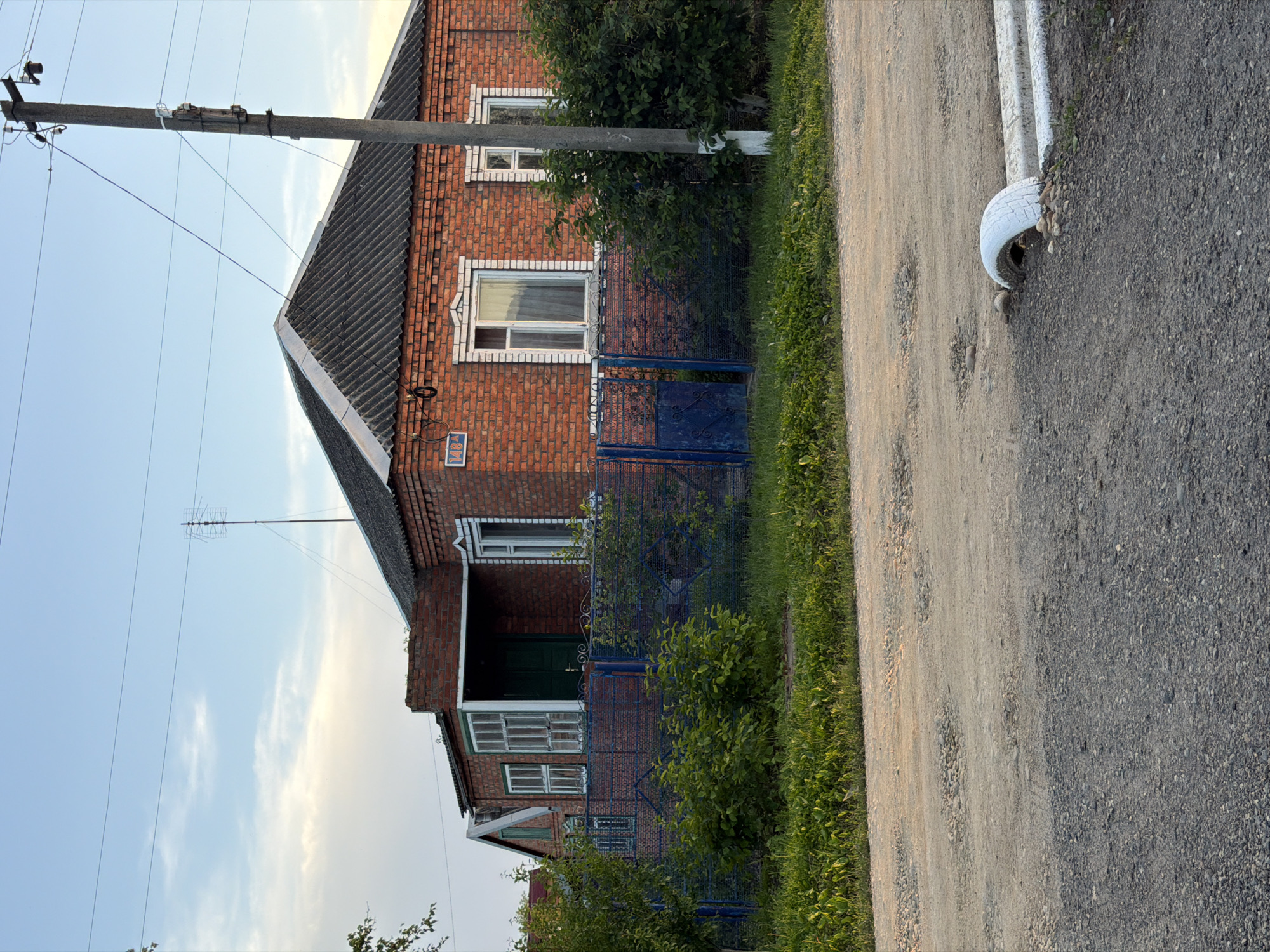 A two-story brick house is adjacent to a gravel road in Leningradskaya, Russia. A blue fence separates the house from the road. A utility pole stands to the left of the house. A painted tire is used as a roadside barrier on the right side of the road. The sky is visible in the background.