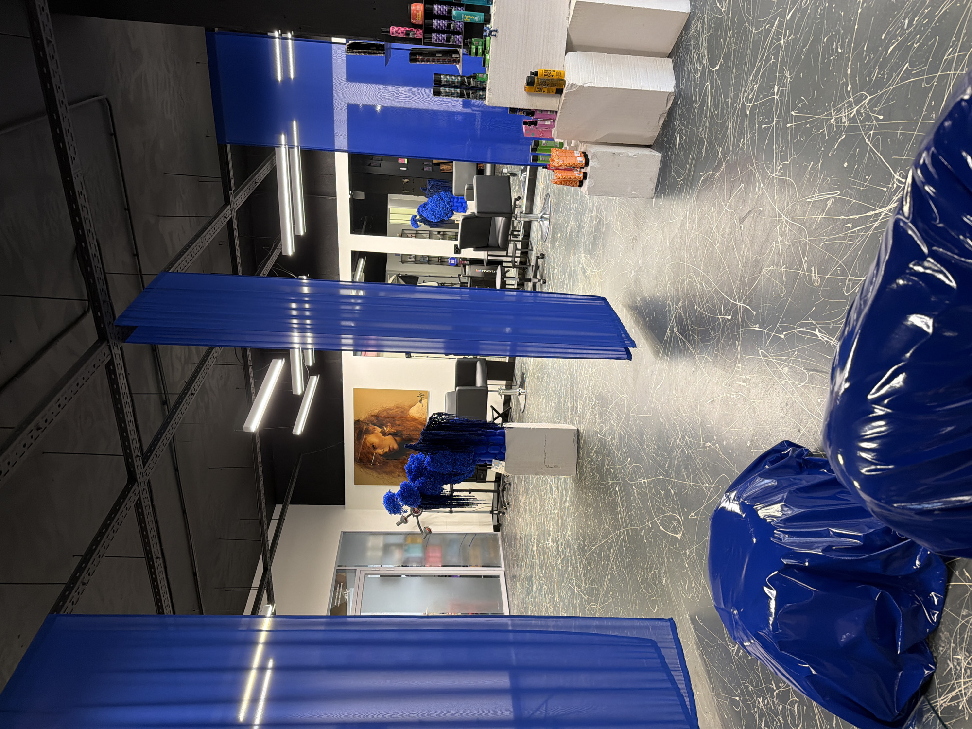 An indoor shot captures a modern salon or studio space. The floor is a dark gray concrete with white paint splatters. Dominating the scene are large, translucent blue fabric panels suspended from the ceiling, creating a vibrant, semi-transparent barrier. In the foreground, two large objects are covered in shiny blue plastic sheeting. A white pedestal in the mid-ground displays a dramatic blue floral arrangement with fringe-like strands. Behind this, a large framed portrait of a woman with fiery red hair hangs on the wall. The space features industrial-style lighting and exposed ceiling infrastructure. Hair salon chairs and mirrors are visible in the background, along with shelves stocked with colorful hair products. The overall aesthetic is contemporary and artistic. The location is identified as a salon in Saint Petersburg, Russia.