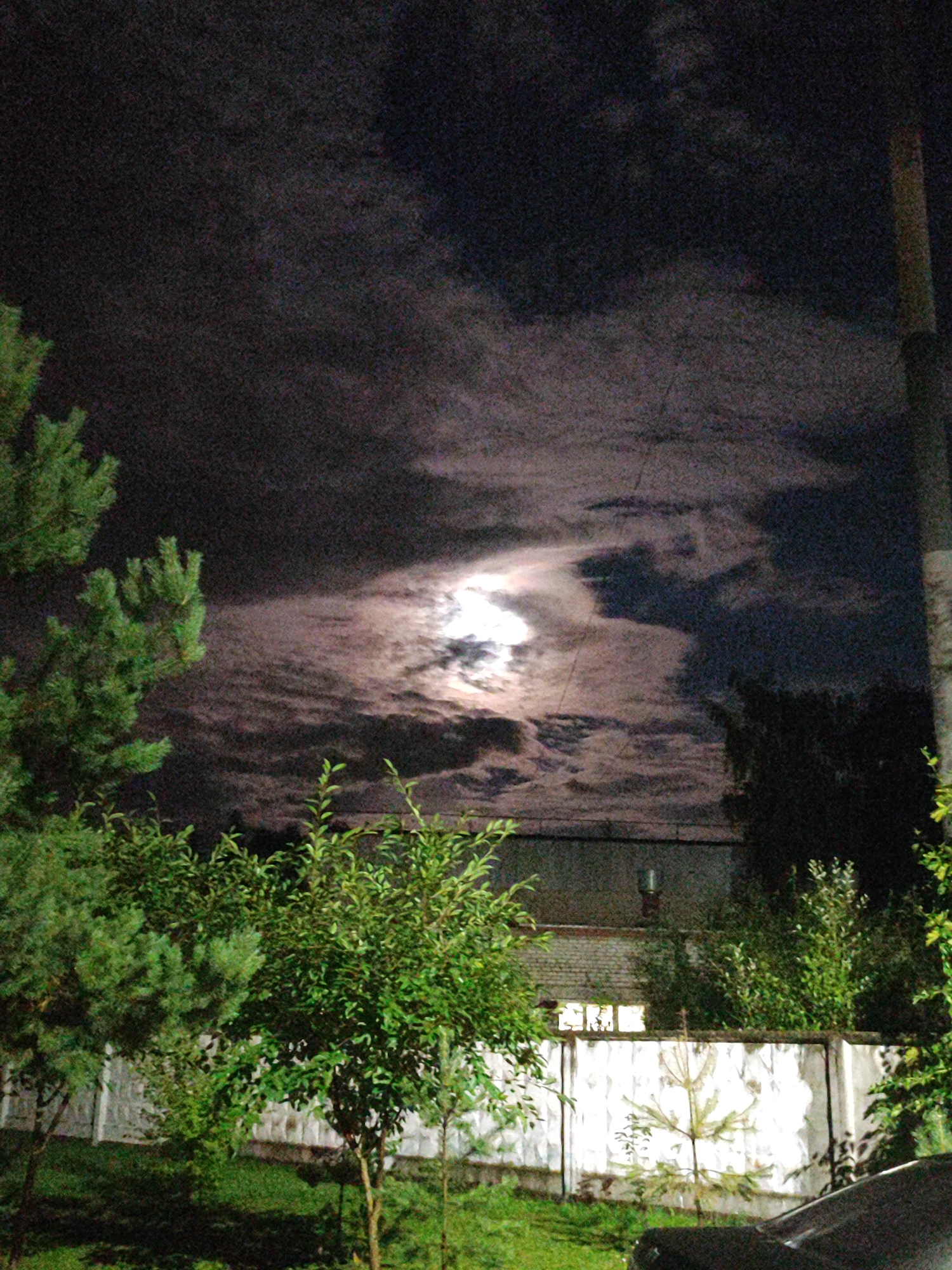 This is a nighttime photograph taken in Bronnitsy, Russia, featuring a cloudy sky with a bright moon visible through a break in the clouds. In the foreground, there are several trees and bushes, with a concrete fence in the midground. A portion of a dark-colored car is visible in the lower right corner of the frame. Behind the fence, a building with windows is partially obscured by vegetation. The overall scene depicts a residential or industrial area at night, illuminated by moonlight and possibly artificial lighting.